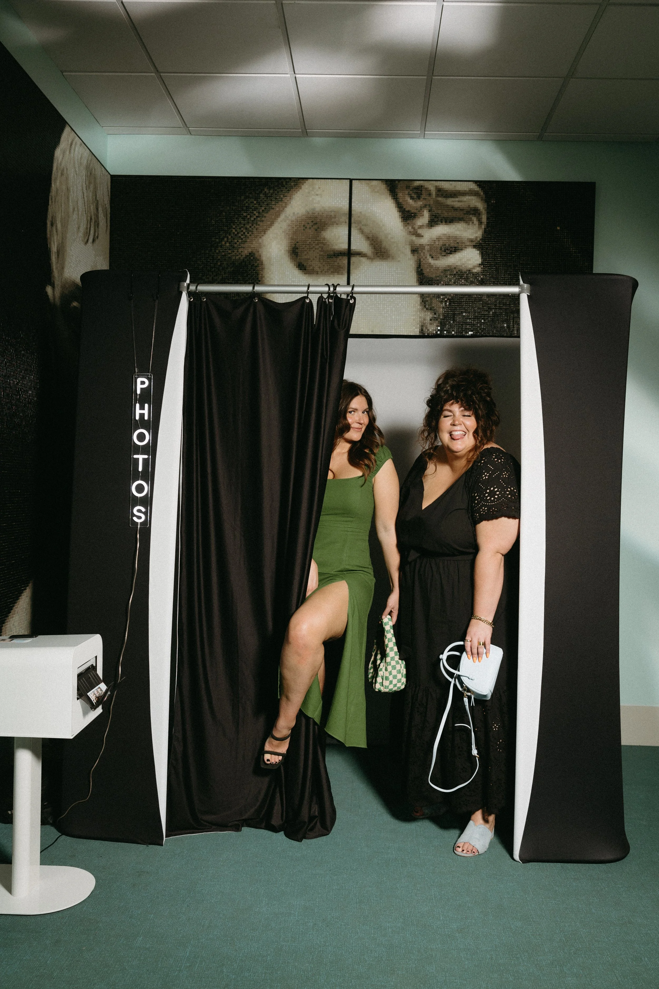 Two women posing in a photo booth, one wearing a green dress with her leg raised and the other wearing a black dress, both smiling and making silly faces.