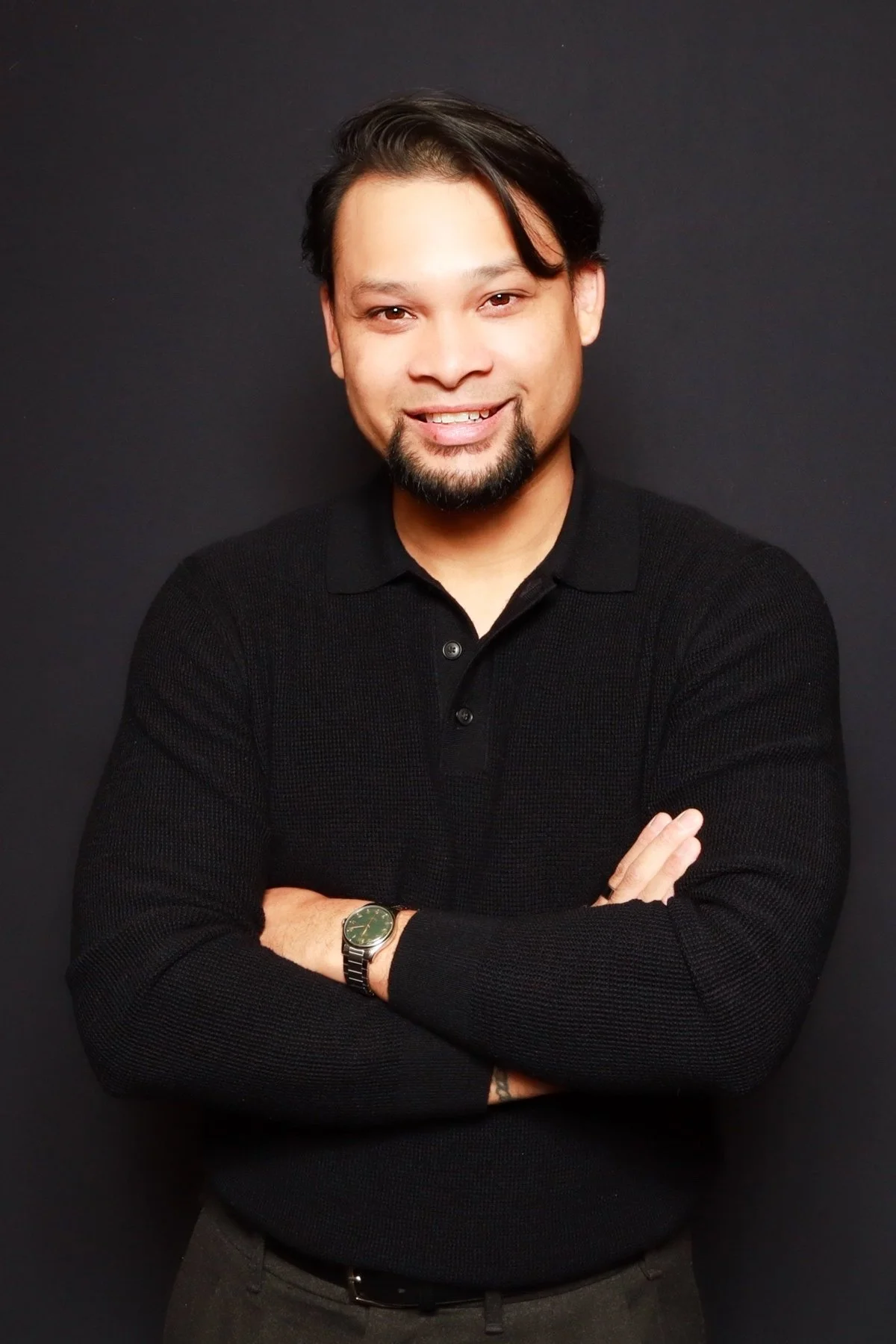 A man with black hair, beard, and mustache is smiling with arms crossed, wearing a black sweater and a watch, standing against a dark background.
