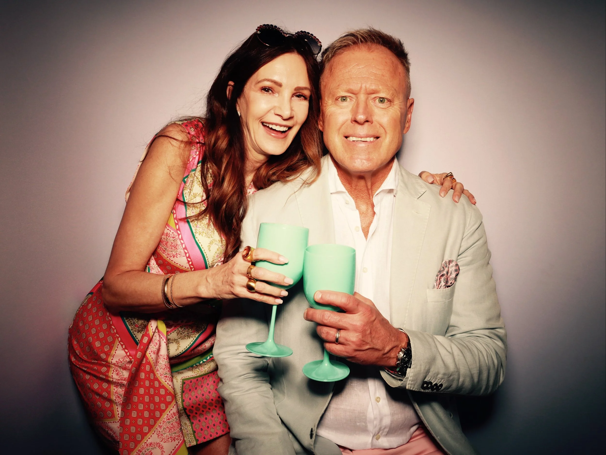 A woman with long brown hair, wearing sunglasses on her head and a colorful patterned dress, is smiling and hugging a man. The man has short blond hair, is wearing a light-colored blazer over a white shirt, and is holding a turquoise champagne flute.