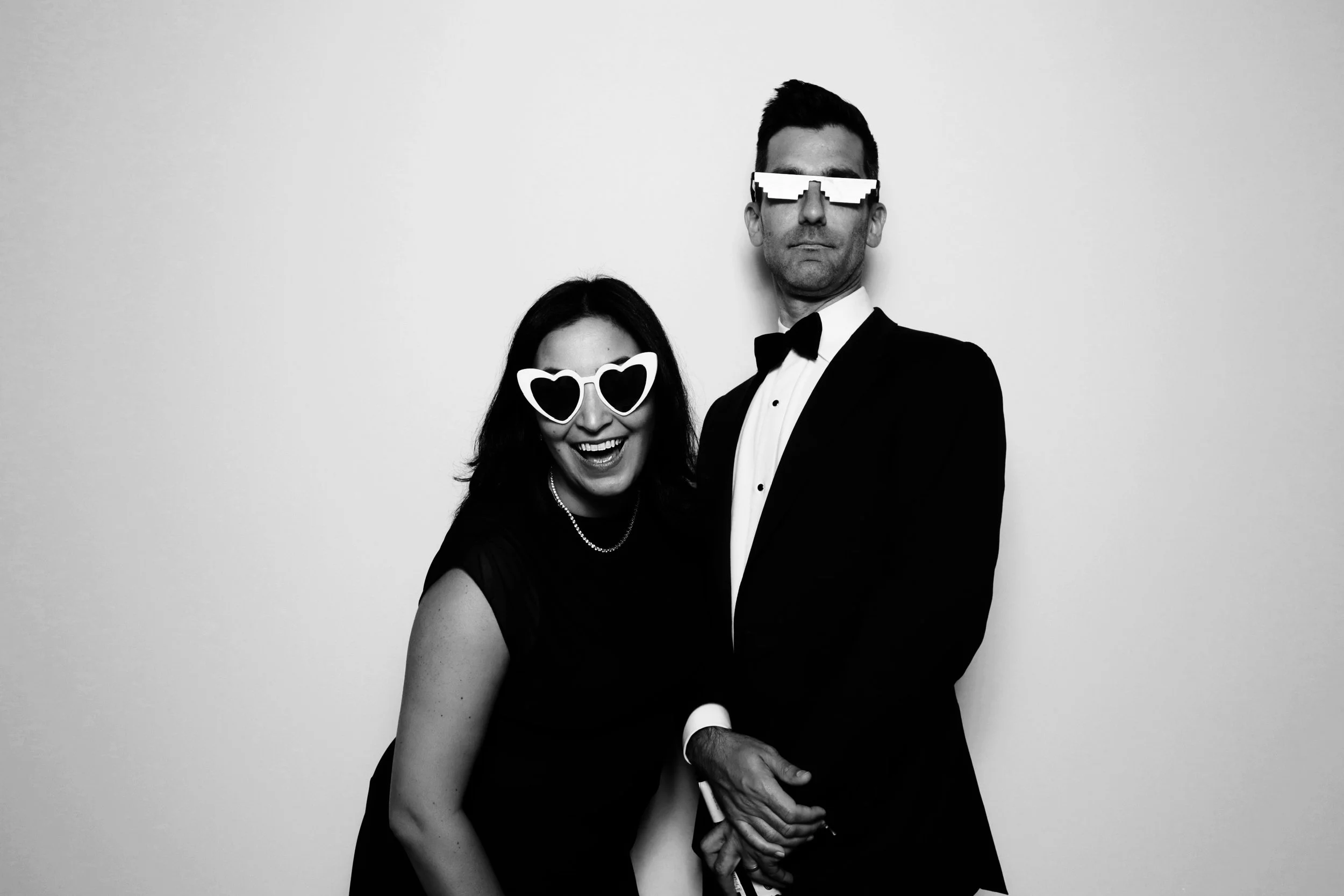 Man and woman dressed in formal tuxedo and evening gown wearing fun sunglasses, posing for a black-and-white photo.