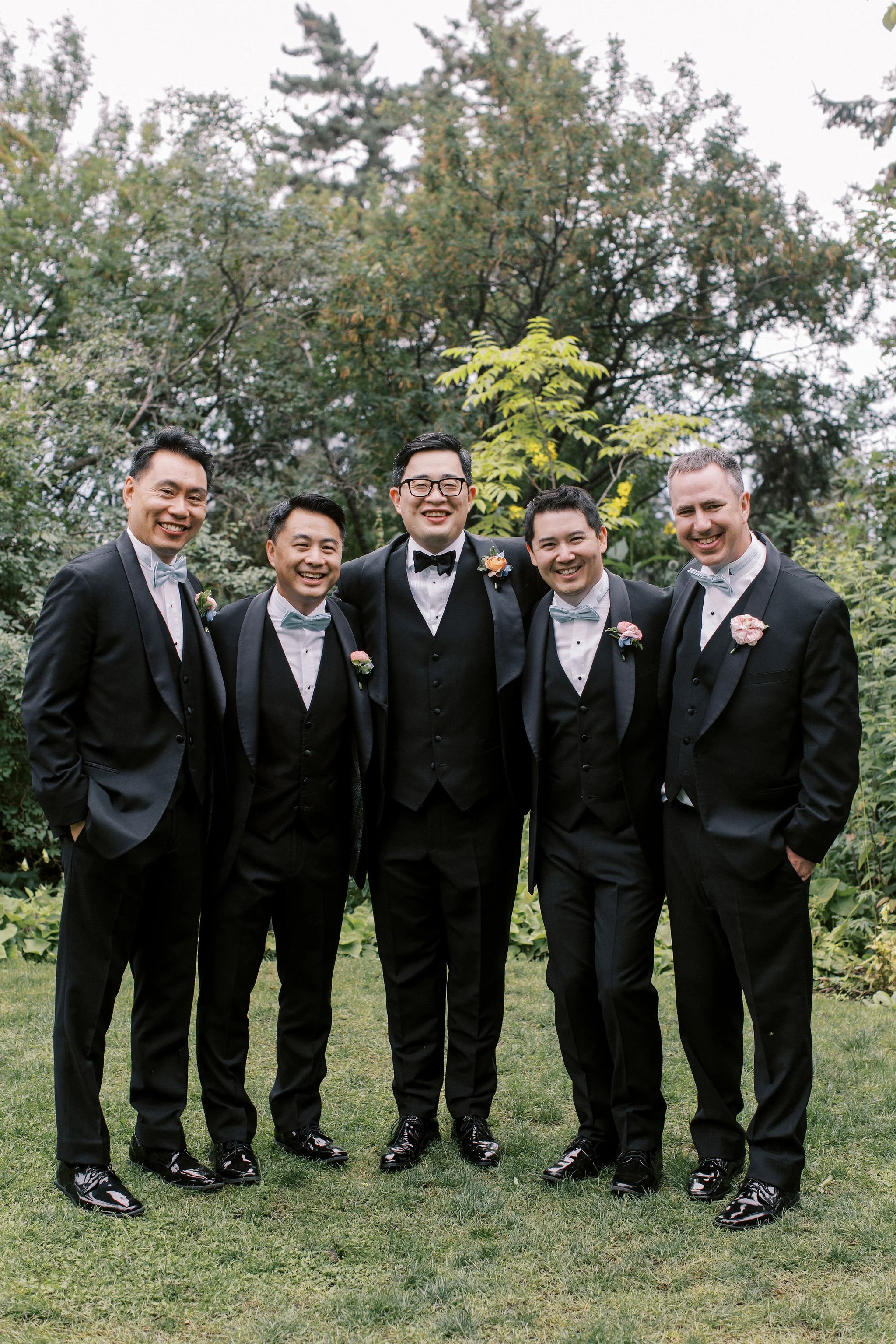 Group of five men dressed in formal tuxedos with bow ties, standing outdoors on grass, smiling, with trees in the background.