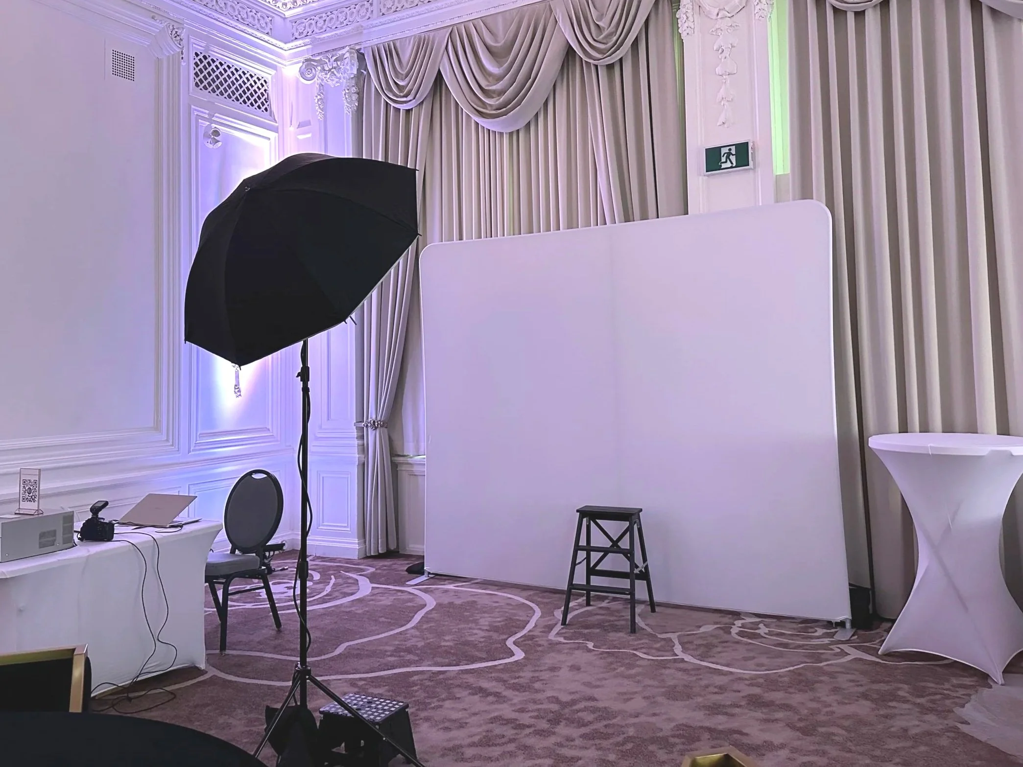Empty event photo booth setup with white backdrop, black stool, photo equipment, and umbrella lighting in a decorated venue.