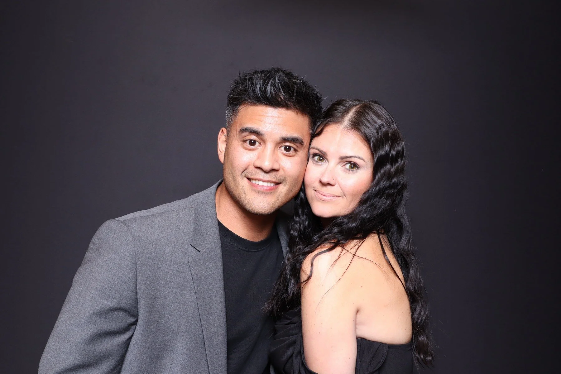 A smiling man and woman posing together against a dark background, with the man wearing a gray blazer and the woman in a black off-the-shoulder top.