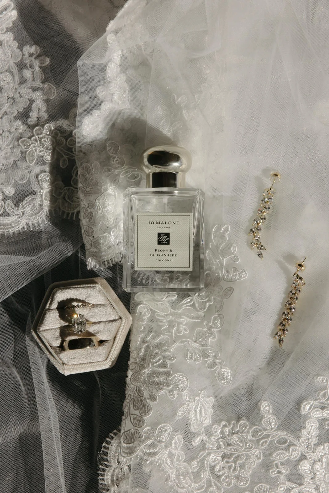 A bottle of Jo Malone Peony & Blush Suede cologne, a jewelry box with an engagement ring and wedding band, and a pair of dangling earrings with crystals, all arranged on white embroidered fabric and lace.