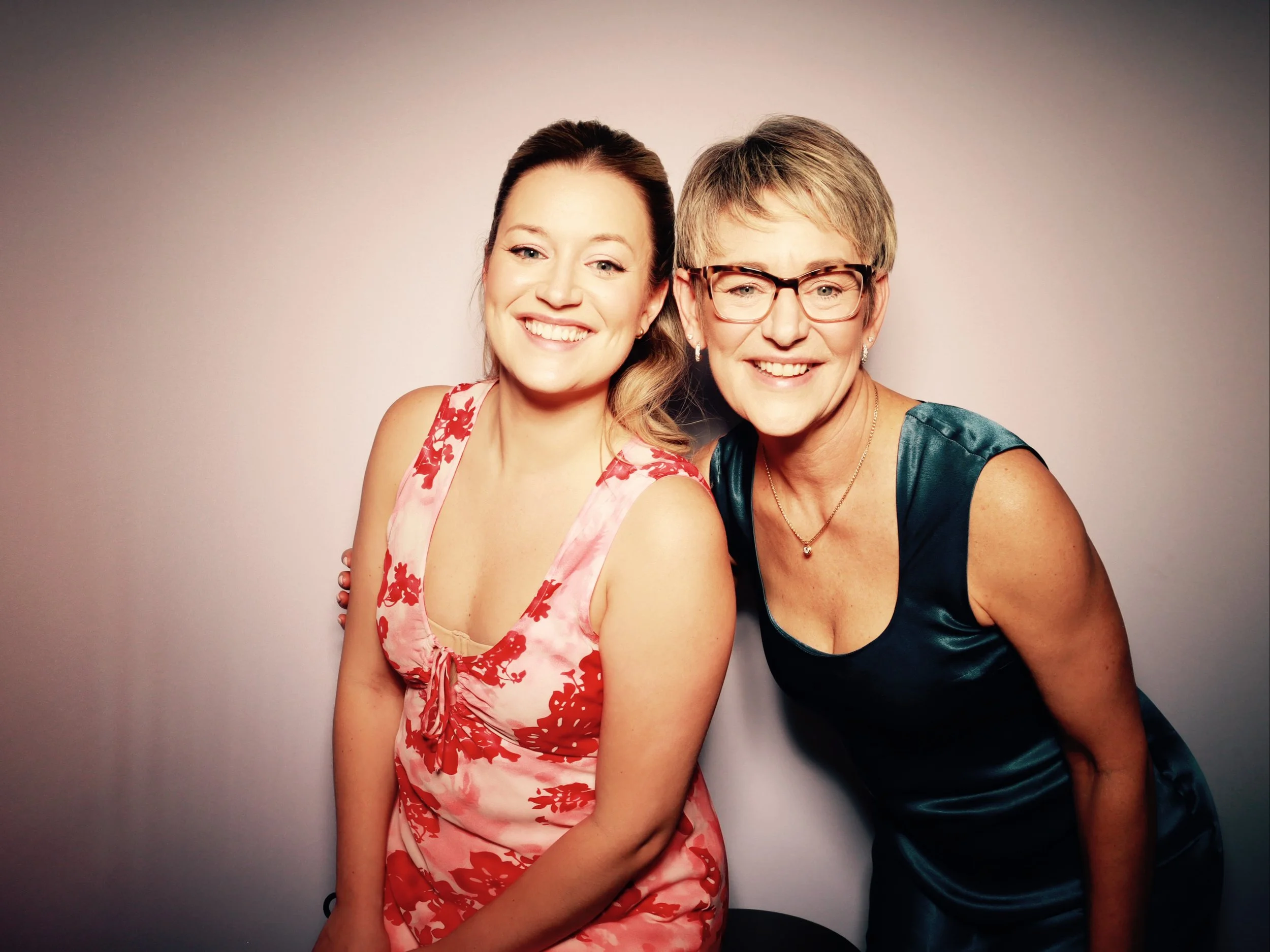 Two women smiling, standing close together against a plain background. The woman on the left has long, wavy hair, and is wearing a sleeveless dress with red floral pattern. The woman on the right has short hair, glasses, and is wearing a dark teal sl