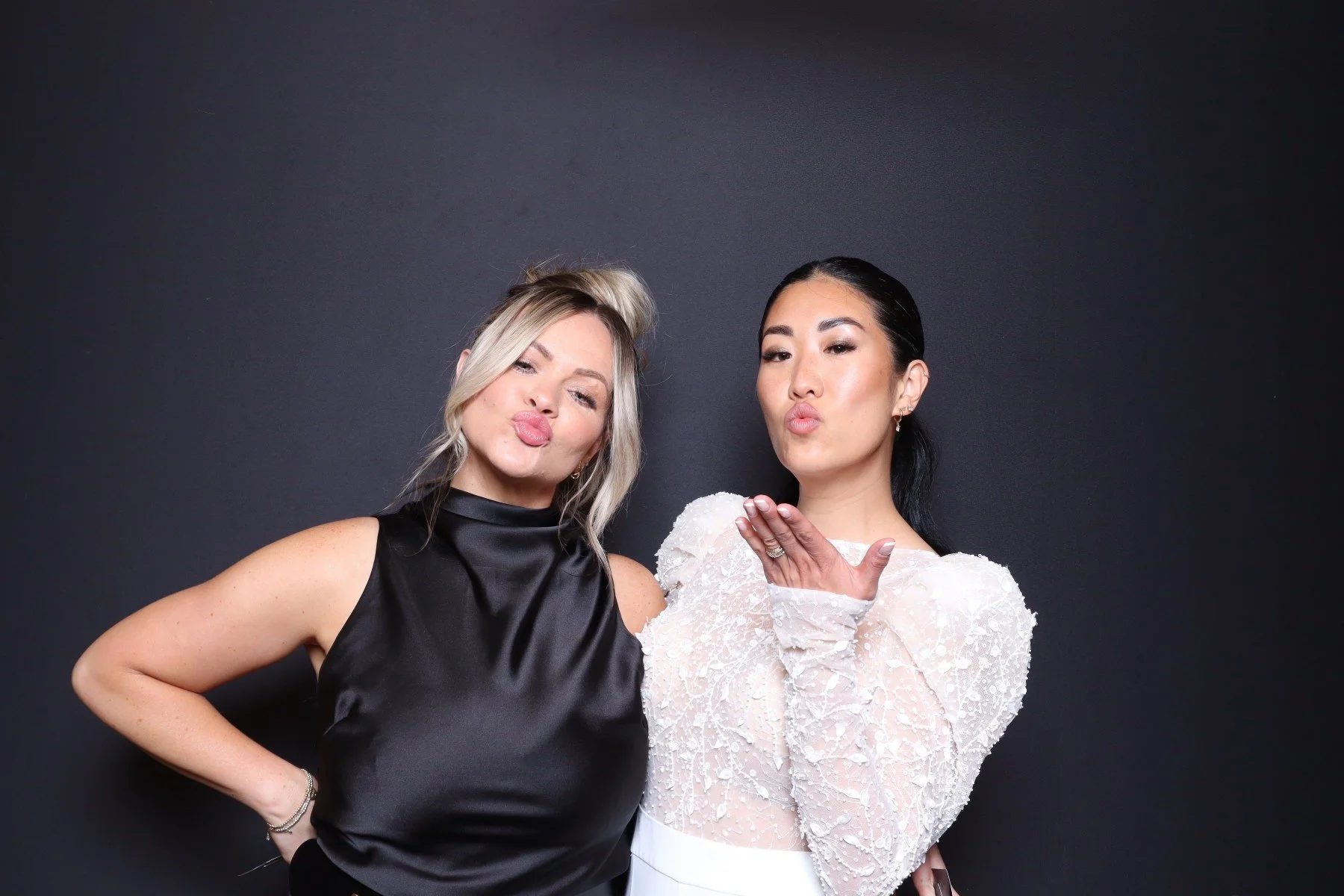Two women blowing kisses at the camera, standing against a black background.