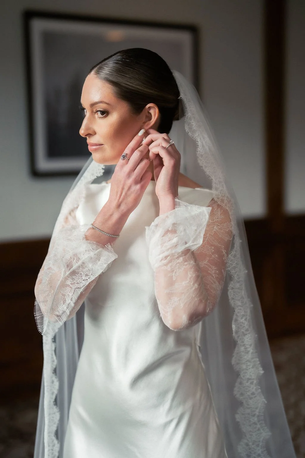 A bride with dark hair styled back, wearing a satin wedding gown with lace long sleeves, is putting on an earring. She has a veil with lace trim over her head and is standing indoors in front of a framed picture.