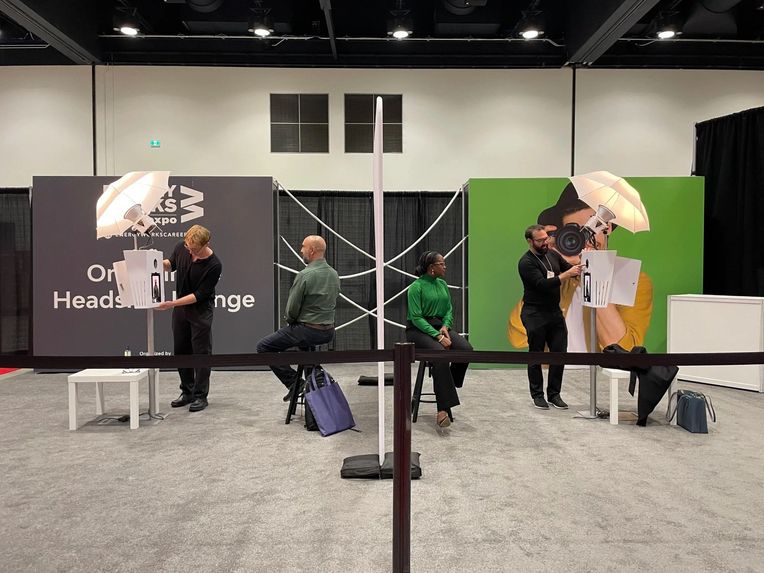 People sitting and standing behind photography booths at an expo, with large camera-themed props and promotional banners in the background.