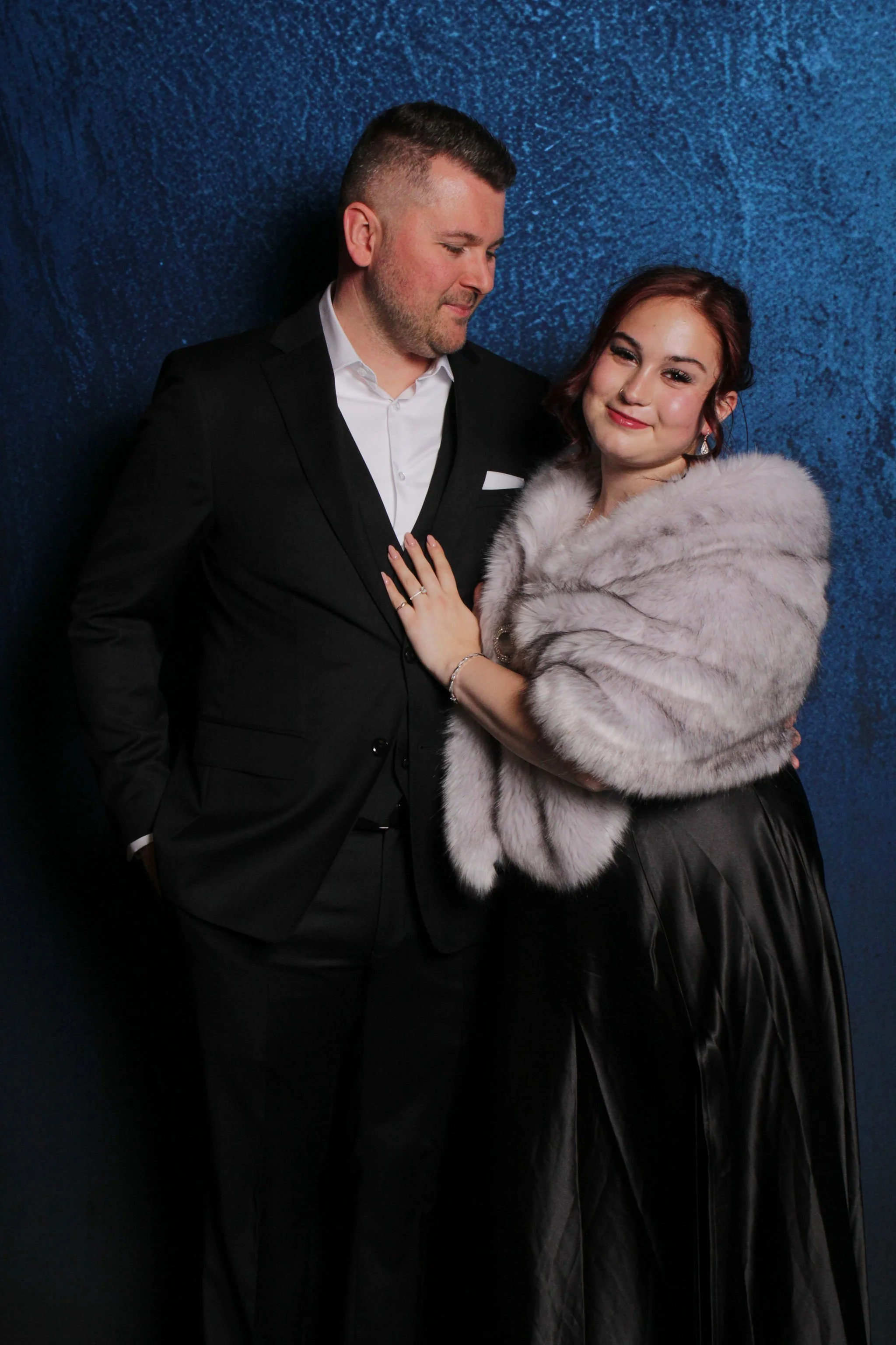 A couple dressed in formal attire stands against a blue textured background. The man, in a black suit and white shirt, is looking down at the woman, who is wearing a faux fur wrap and a black satin skirt. She is smiling and looking at the camera, wit