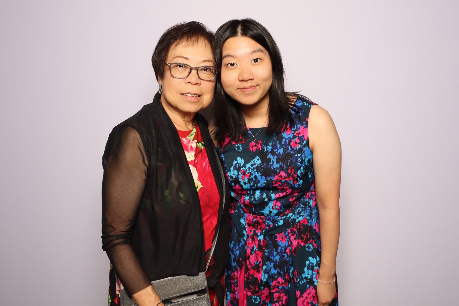 An older woman and a younger woman standing close together, smiling, against a plain light-colored background.
