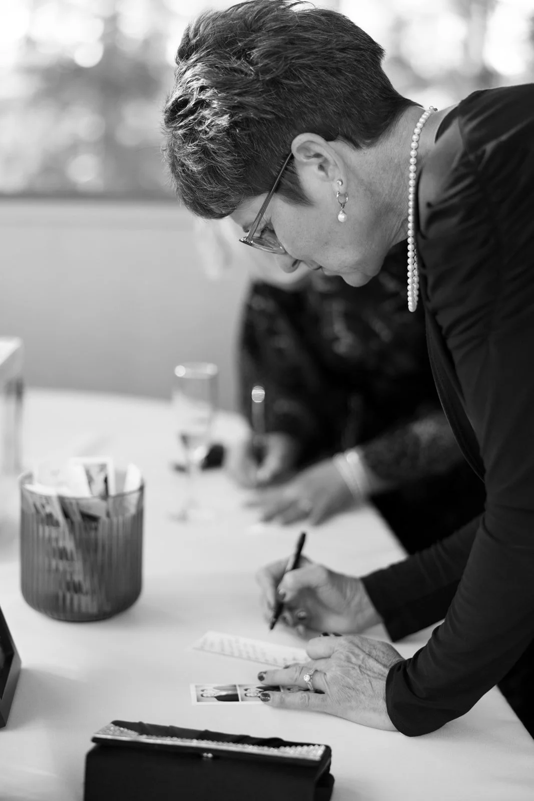 A woman with short hair and glasses signing a document at a table, with a stack of photographs in front of her in an indoor setting.