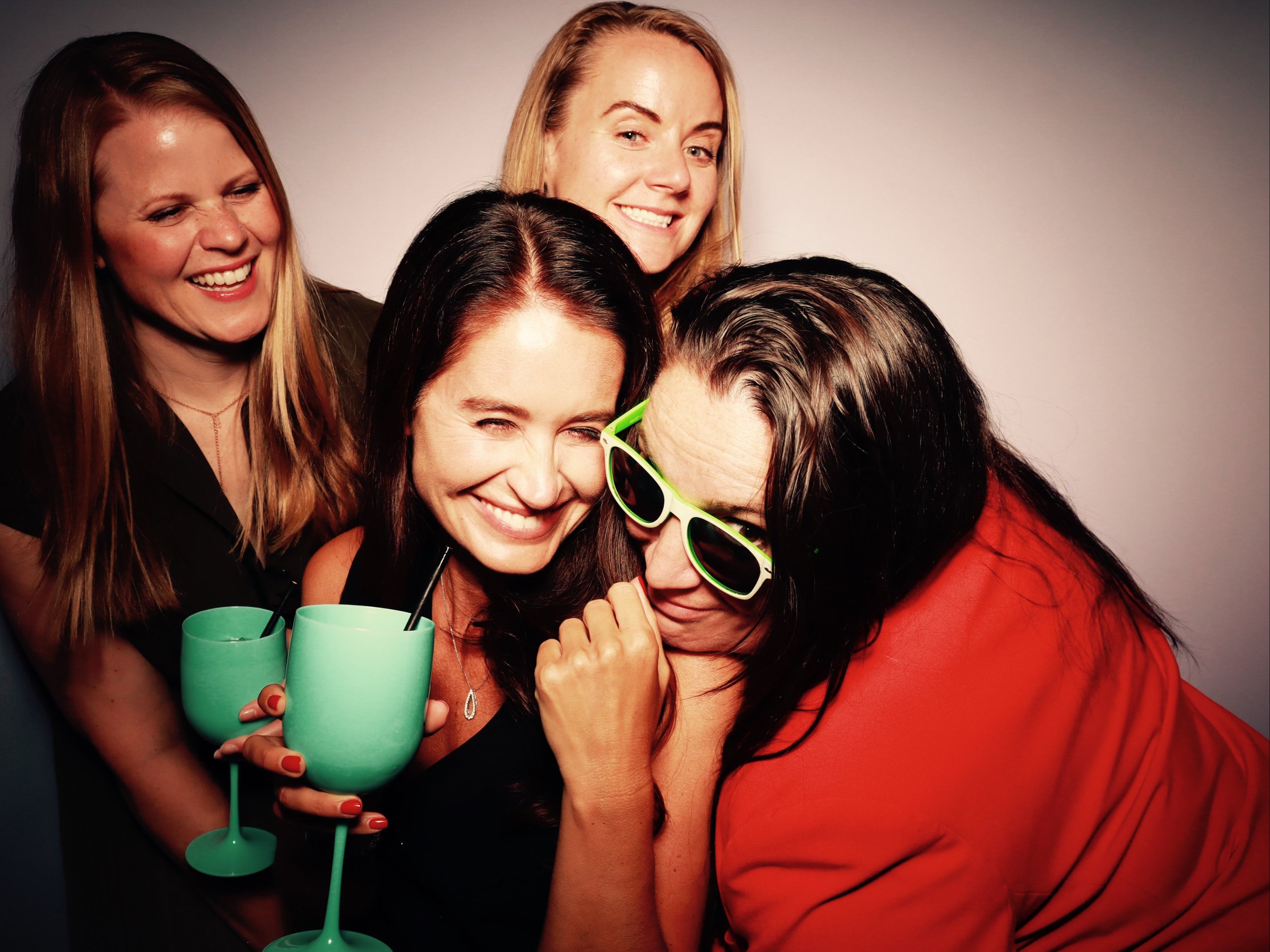 Four women enjoying a celebration with two women holding turquoise drinks and wearing sunglasses, all smiling and having fun together.