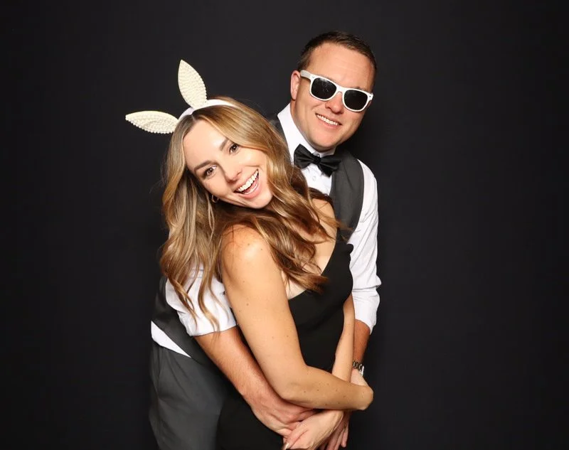 A smiling woman with long, wavy hair wearing bunny ears, and a smiling man wearing sunglasses, a white shirt, and a black vest and bow tie, posing together against a dark background.