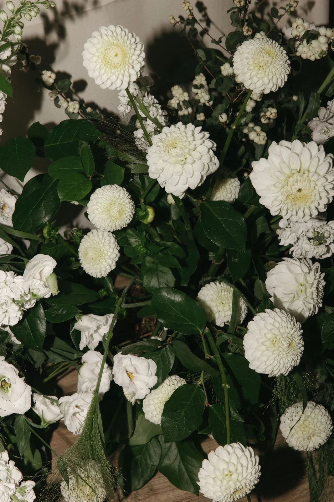 A floral arrangement of white dahlias, white roses, and white berries with green leaves and foliage.