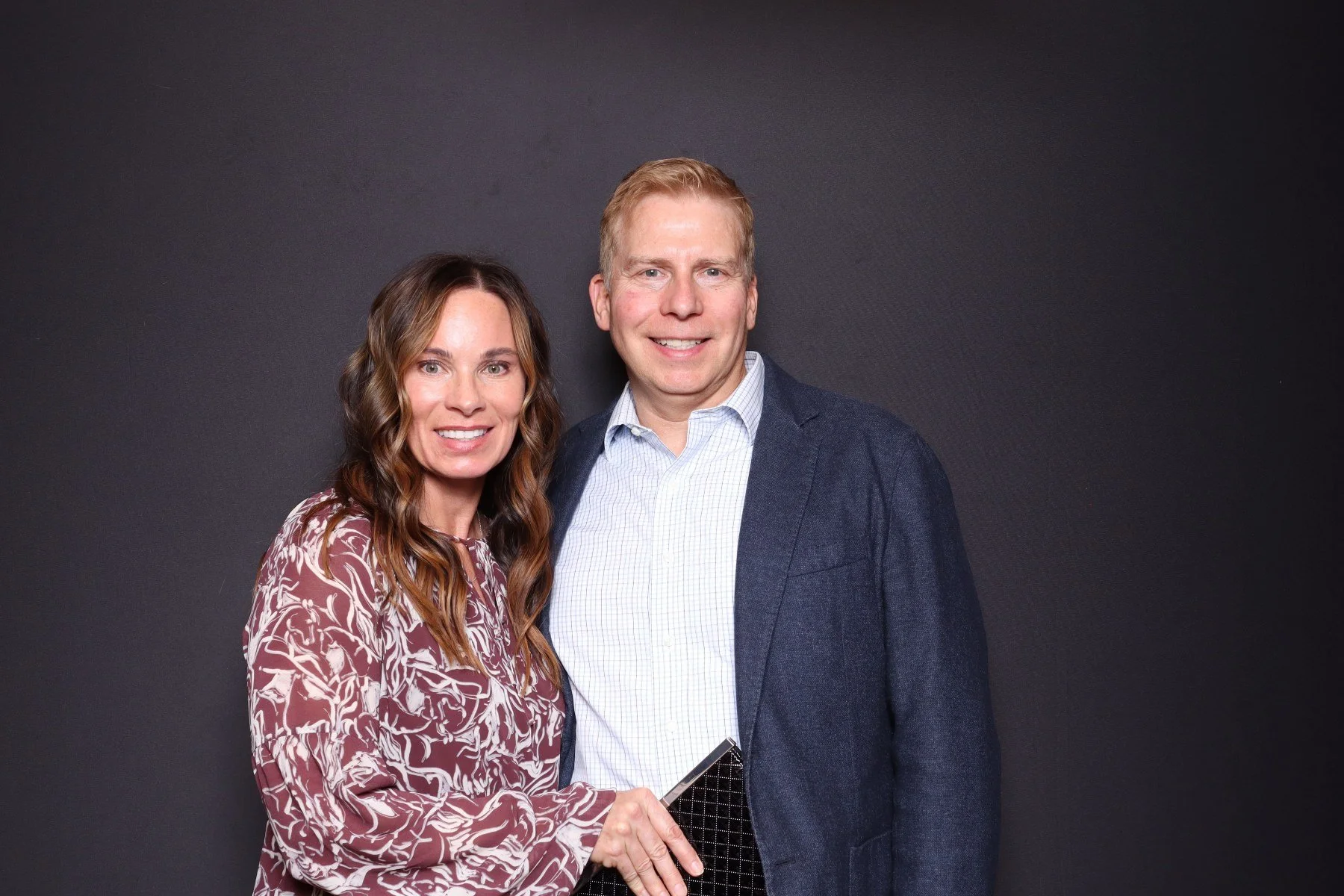 A woman with long wavy brown hair and a man with short light-colored hair smiling and standing close together against a dark background, holding a black craft notebook.
