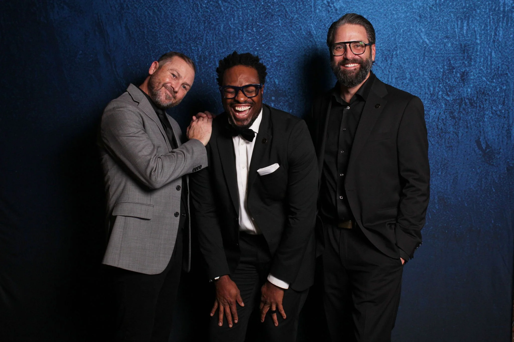 Three men wearing suits and glasses standing and smiling against a dark blue textured background.