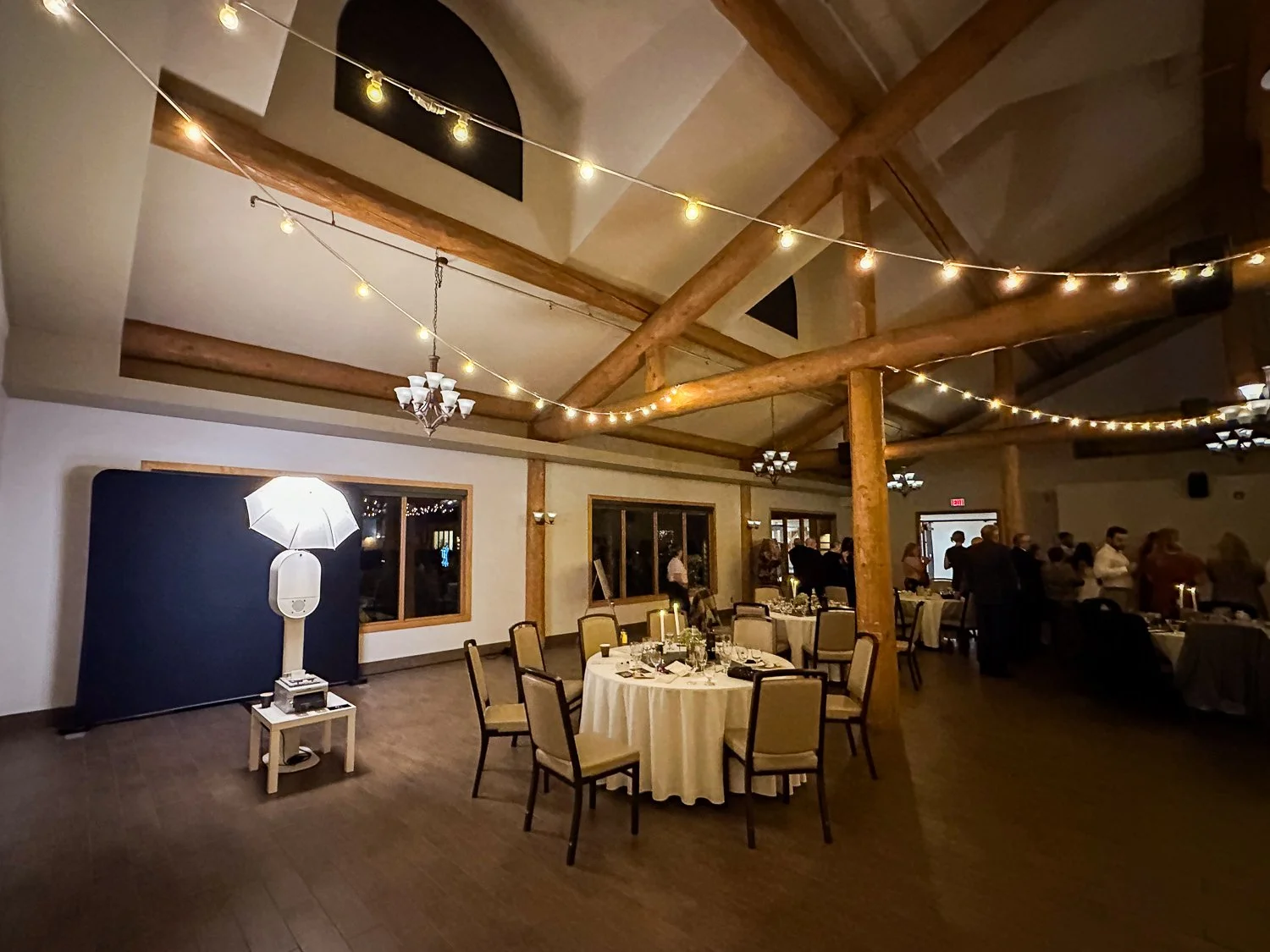 Event hall decorated with string lights, wooden beams, and chandeliers, featuring a table with a beige tablecloth and chairs, with a photo booth setup with a white umbrella at the front.