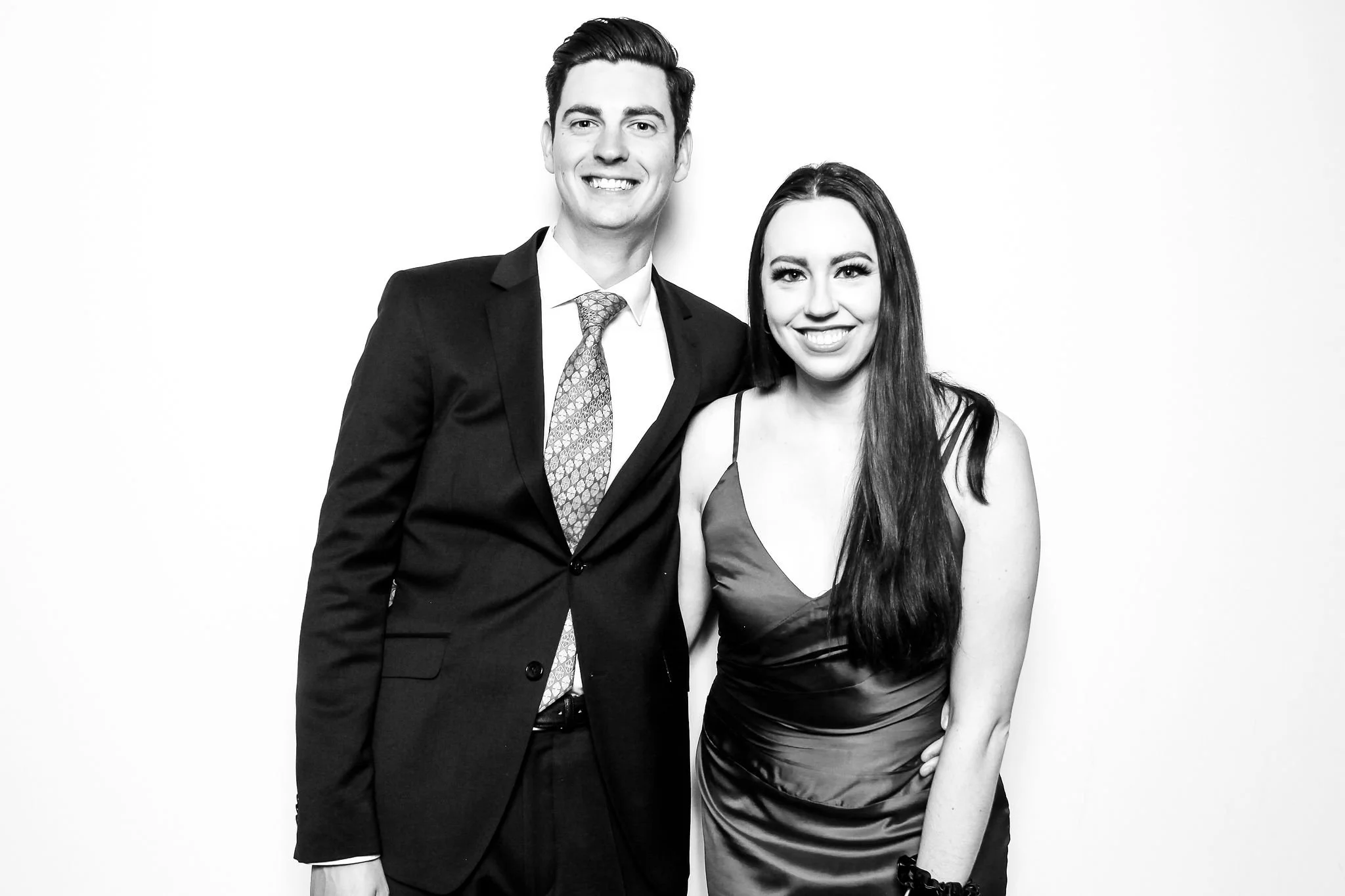 A smiling man and woman dressed in formal attire standing together against a plain background, with the man wearing a suit and tie and the woman in a satin dress.