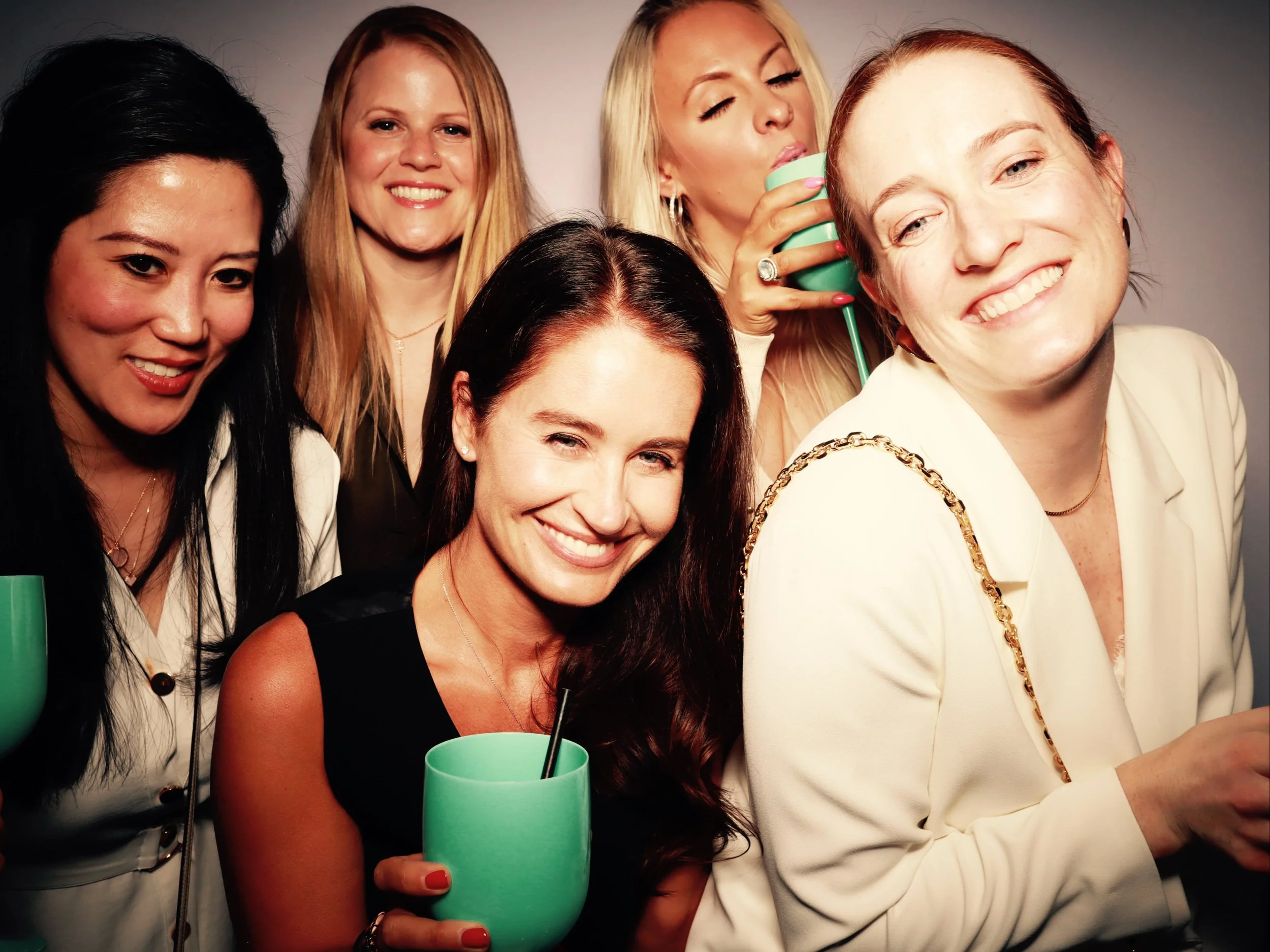 Group of five women enjoying a party, smiling and holding green cups with black straws, dressed stylishly against a plain background.