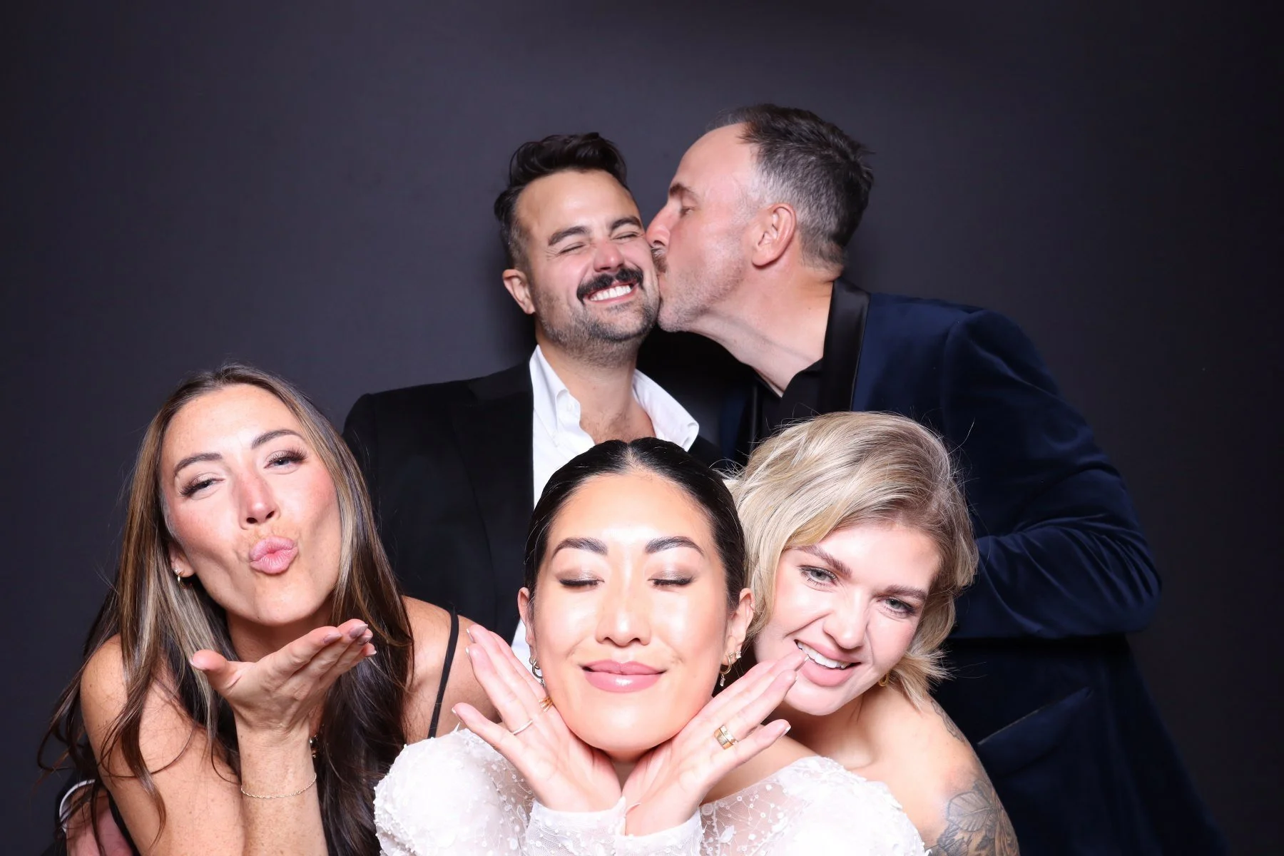 A group of five diverse friends celebrating, with one woman under a veil, one man kissing another on the cheek, and all smiling and making playful gestures against a dark background.
