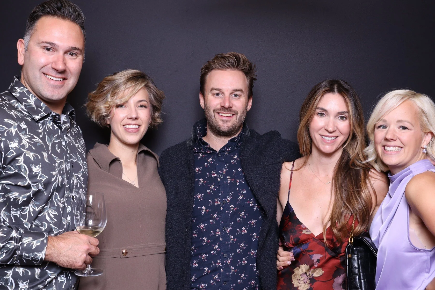 Group of five smiling people, two men and three women, standing together in a party or social gathering, with one woman holding a wine glass.