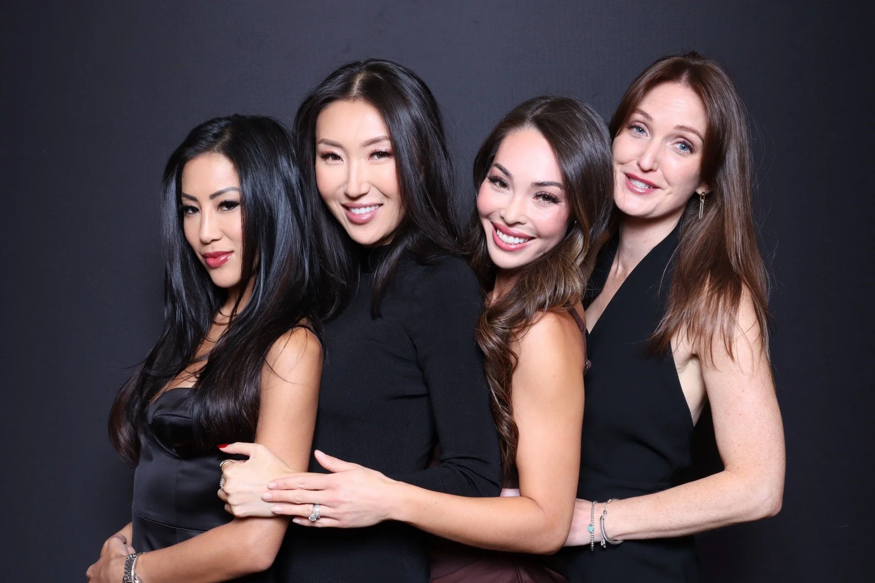 Four women in elegant black dresses smiling and posing together against a dark background.