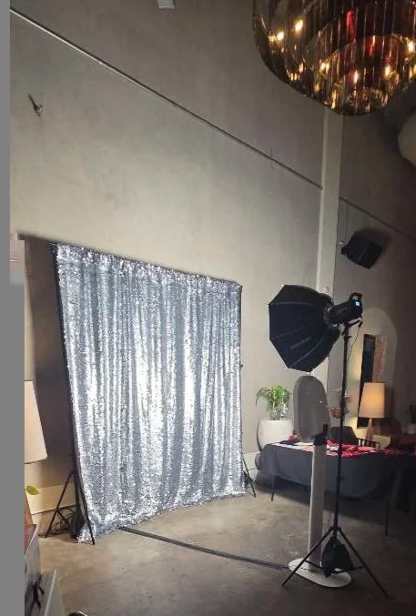Photo of a photography or photo booth setup with a silver sequin backdrop, a studio light with a softbox, and a table with a chair in a room with concrete walls and a chandelier.