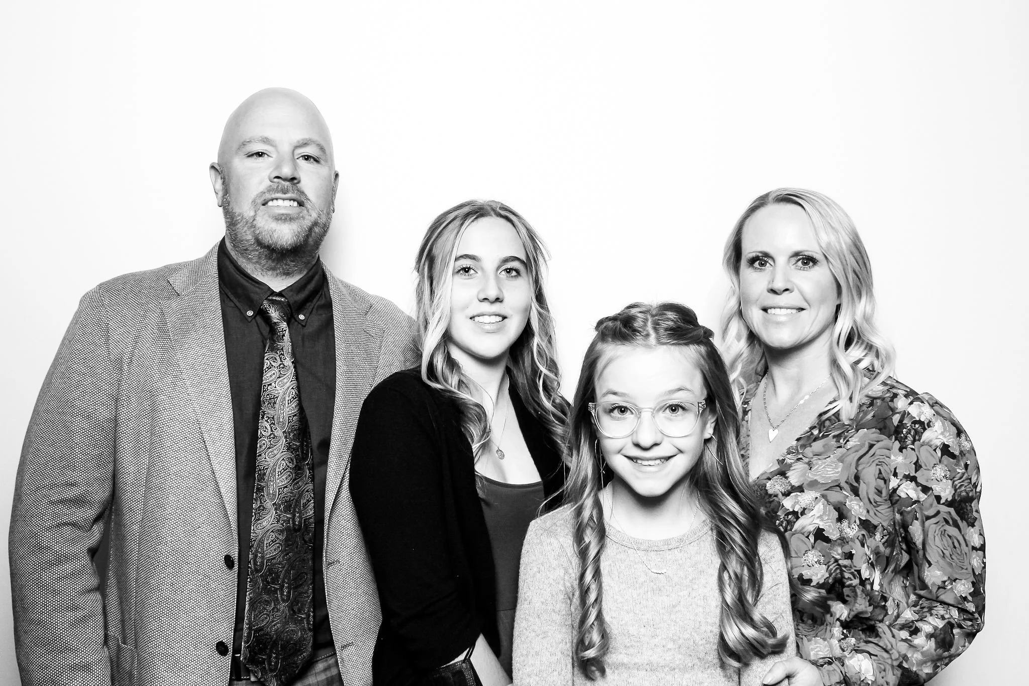 A family of four standing together against a plain background, smiling at the camera. The father is on the far left, the mother on the far right, with two daughters between them. The younger daughter has glasses, and the older daughter has wavy hair.