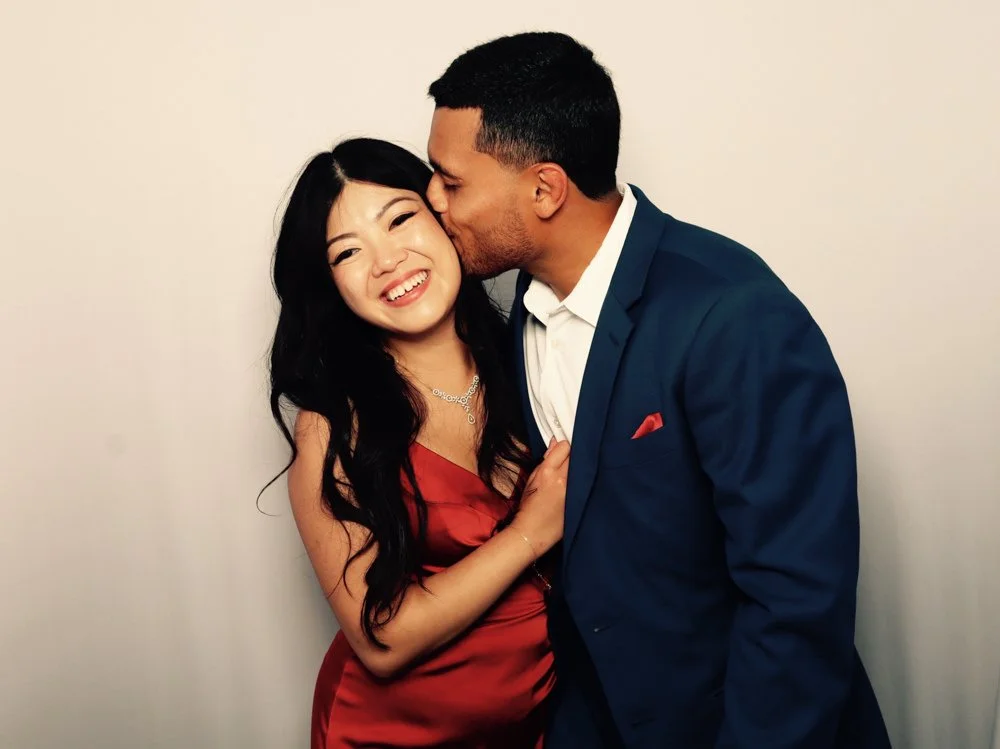 A couple dressed in formal attire, with the man kissing the woman's cheek as she smiles.