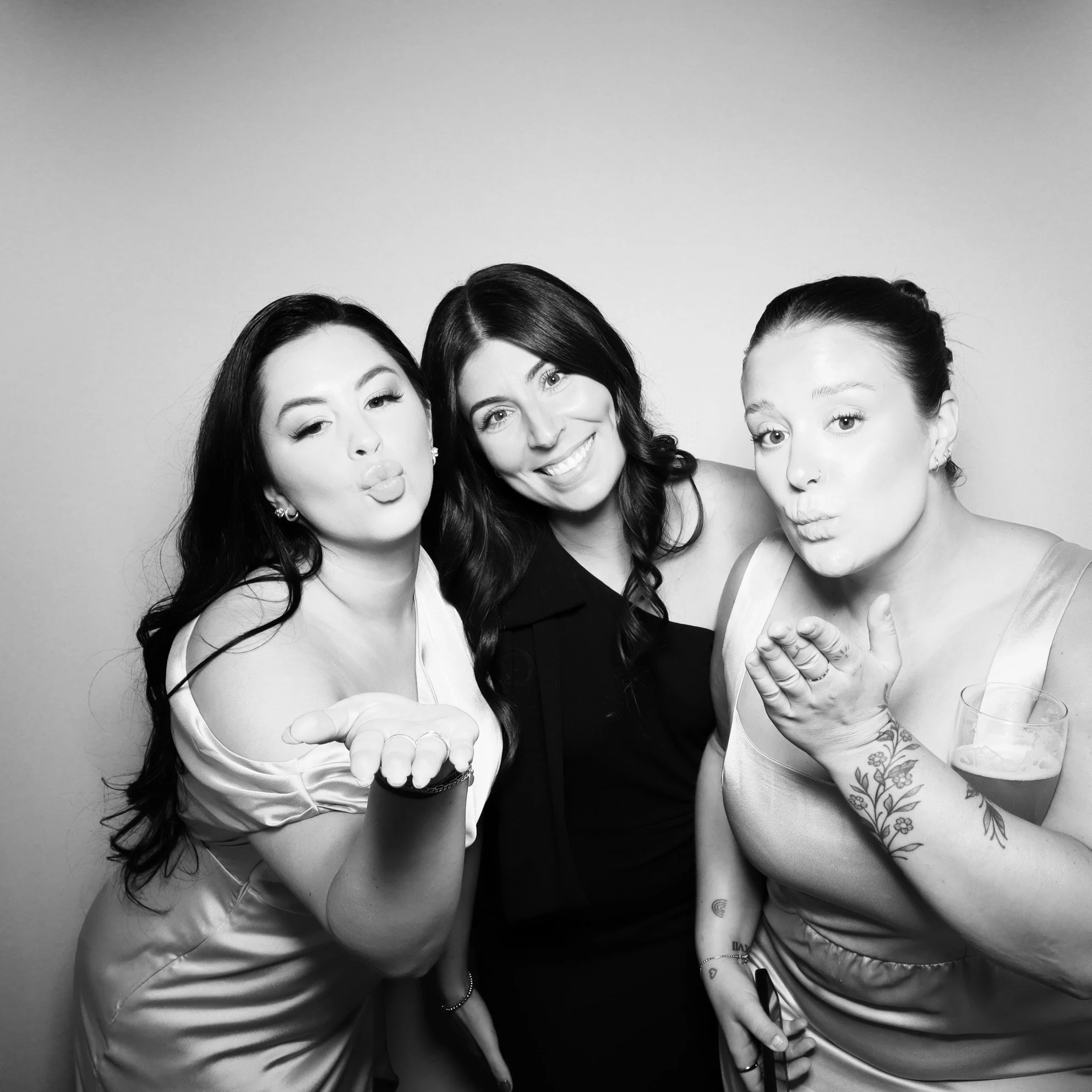 Three women posing together, wearing dresses, some blowing kisses, one holding a drink, in a photo booth setting.