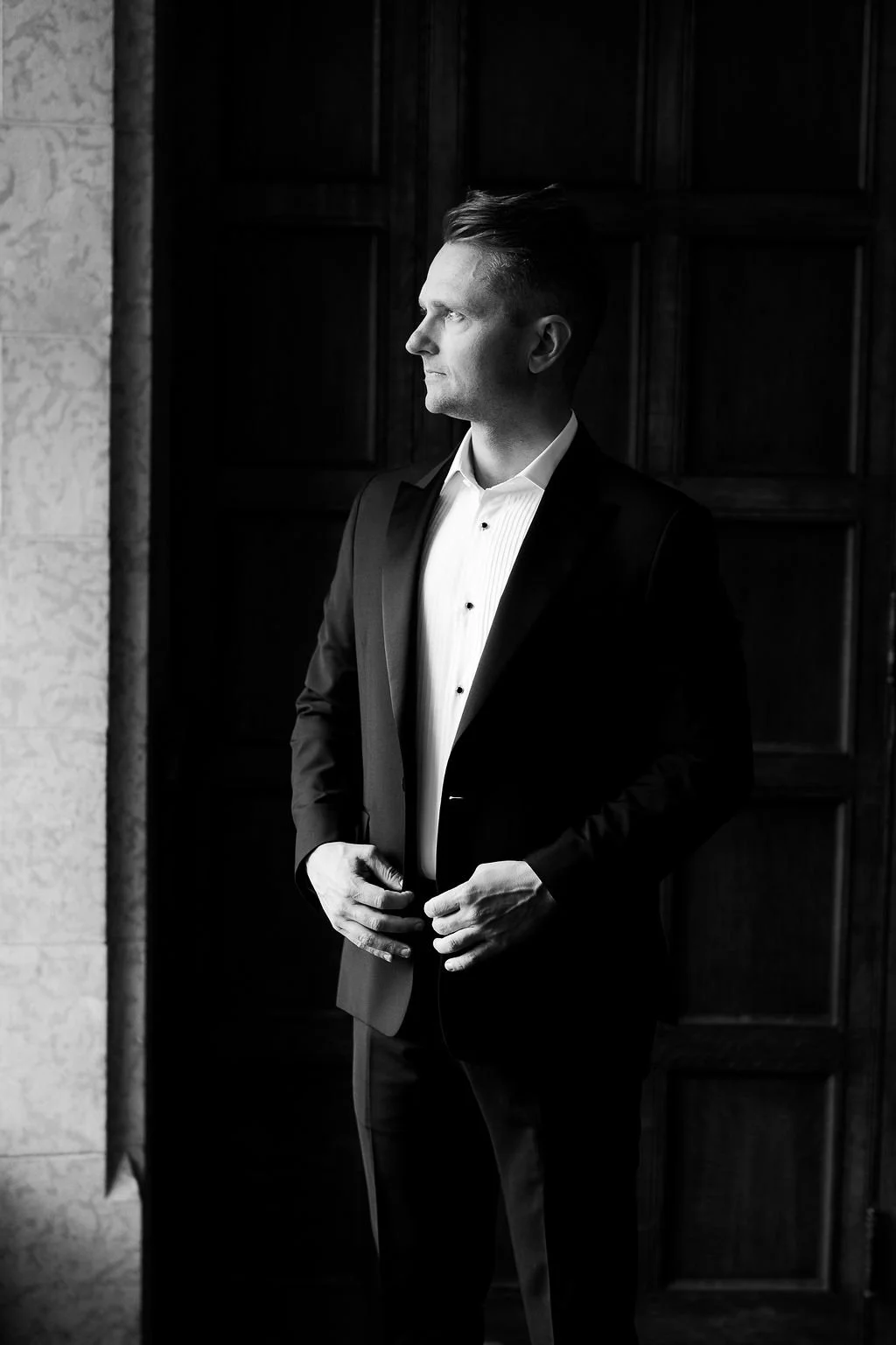 Black and white photo of a man in a tuxedo standing indoors, looking to the left, with wooden paneling in the background.