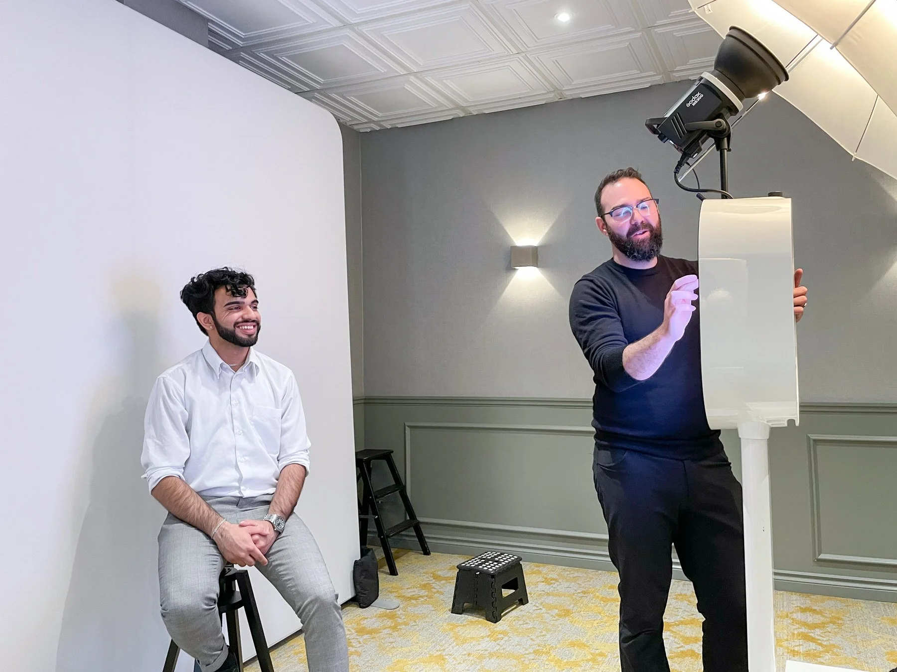 A man with glasses and a beard is operating a photo booth camera, while another man with dark, curly hair, a beard, and a white shirt sits on a stool, smiling during a photo session in a room with gray walls and yellow patterned carpet.