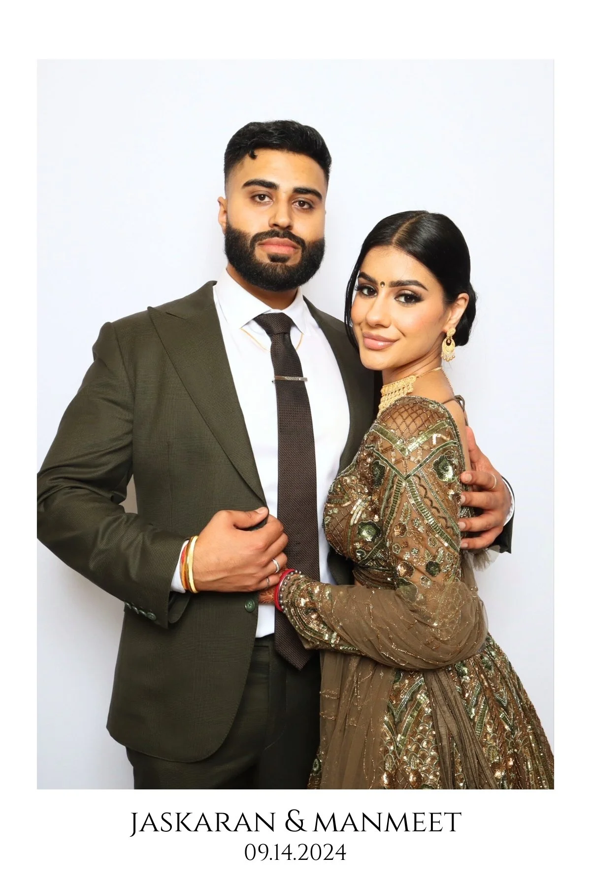 A couple dressed in formal attire, standing close together against a plain white background. The man is wearing a dark green suit with a white shirt and a dark tie, and has a well-groomed beard and hairstyle. The woman is wearing a heavily embroidered gold and brown traditional dress, with jewelry including earrings, a necklace, and bangles. They are holding hands, and the image is captioned with their names