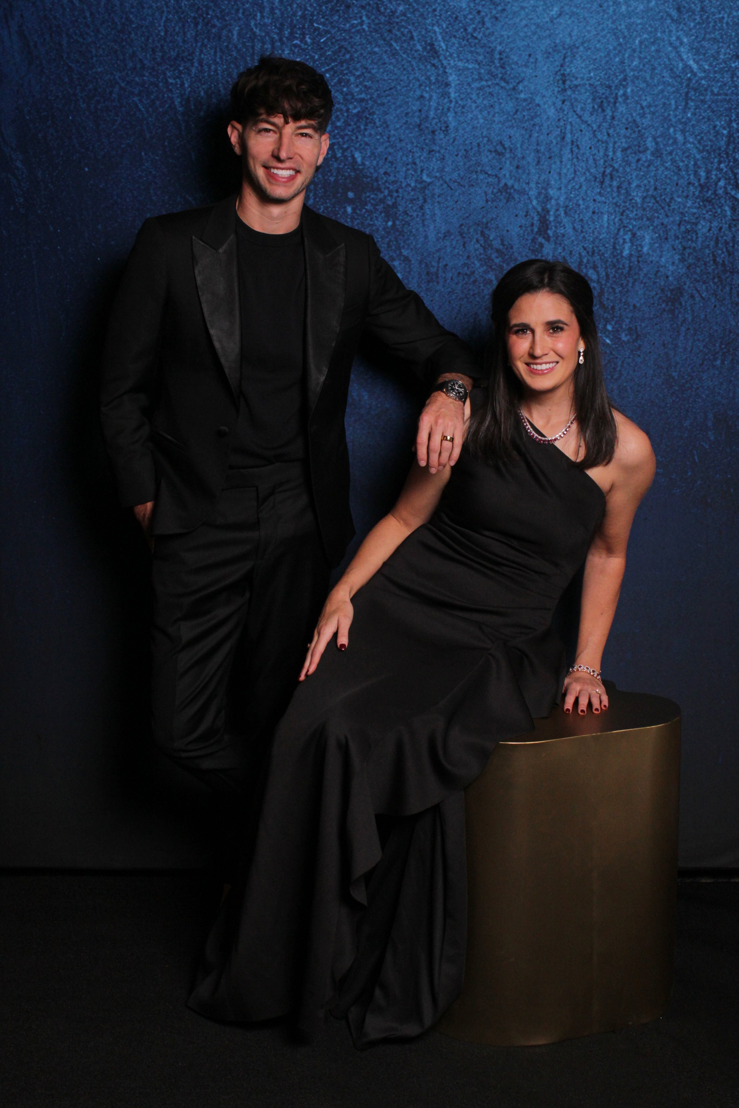 A man and woman in formal black attire posing together against a textured dark blue background. The woman is sitting on a gold cylindrical stool, and the man is standing beside her with one hand resting on her shoulder. They are smiling and looking a
