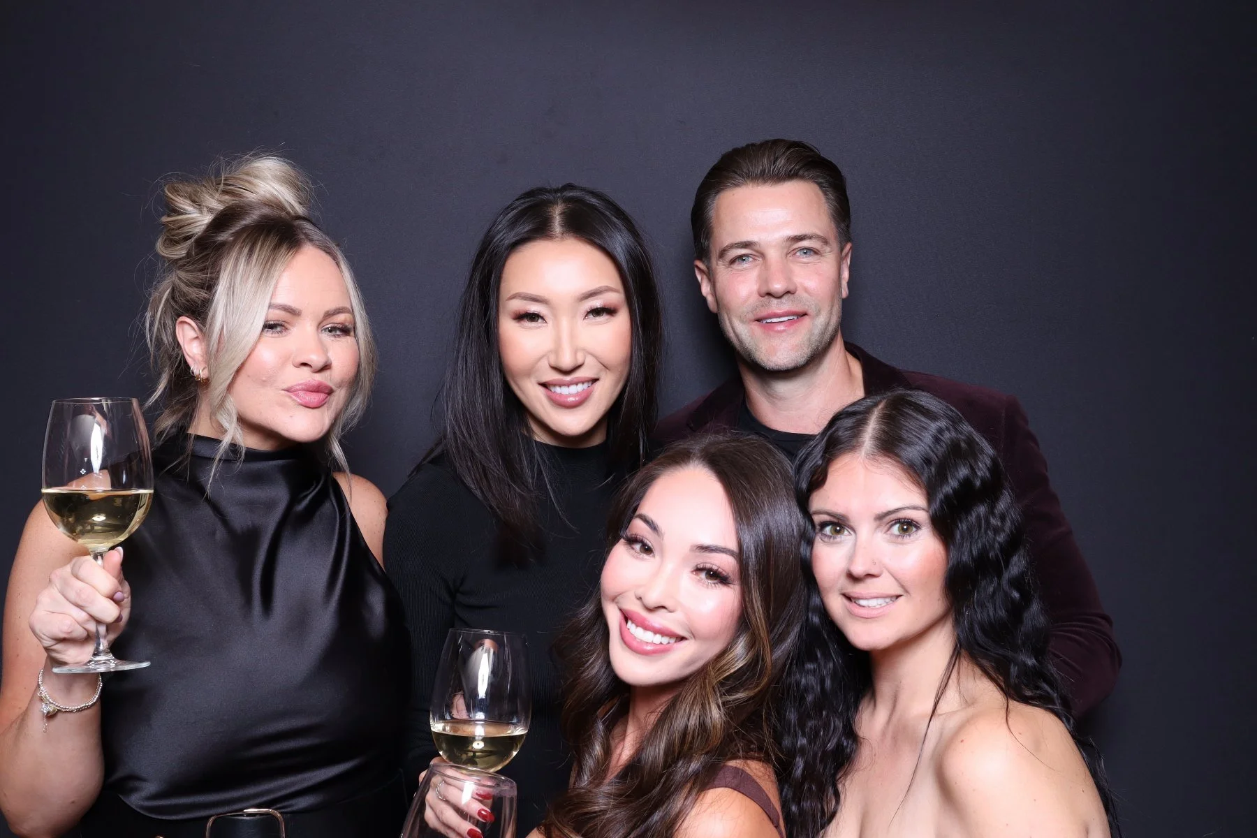 Group of five adults at a party, holding glasses of white wine, smiling against a dark background.