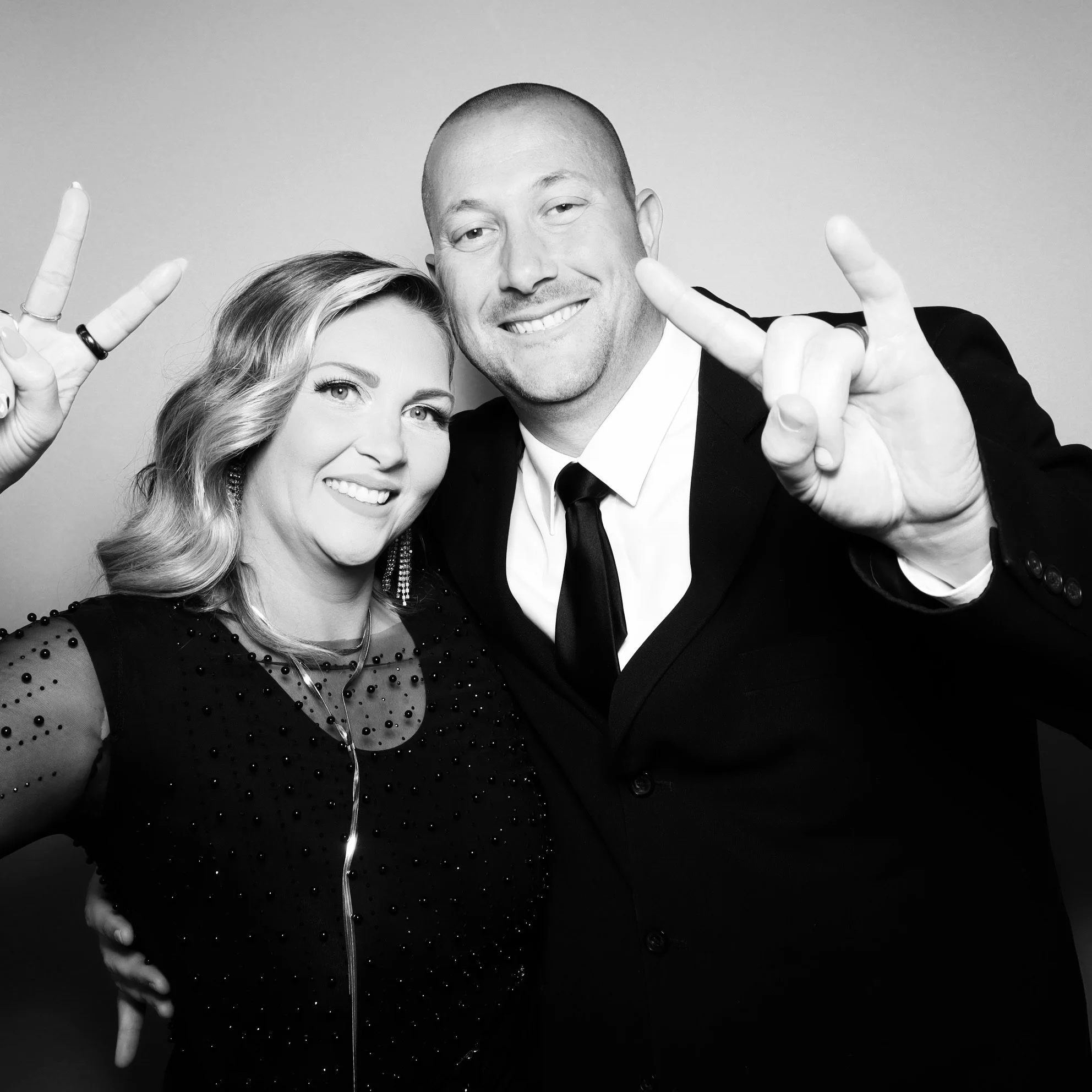 A man and woman posing for a photo, both making peace signs with their right hands. The woman has wavy hair and is wearing a black dress with embellishments, while the man is dressed in a suit and tie. They are smiling and standing close together.