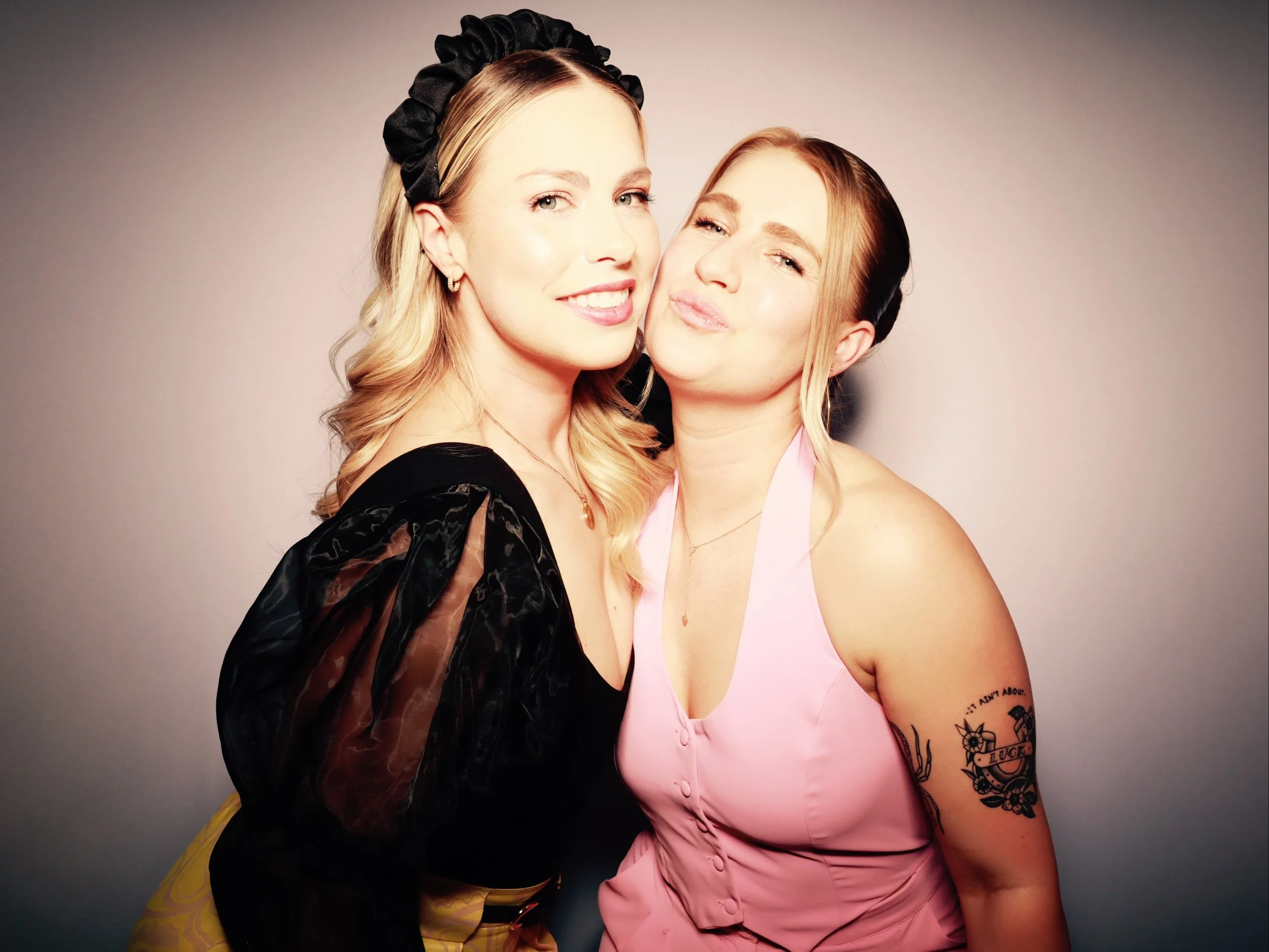 Two women smiling and posing closely with a neutral background. The woman on the left has blonde hair, a black headband, and wears a black dress with sheer sleeves. The woman on the right has reddish-brown hair, is wearing a pink halter top, and has 
