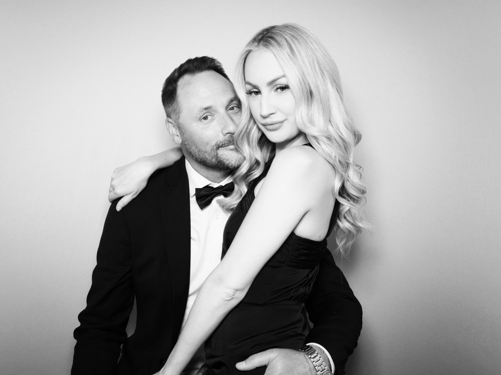A black and white photo of a man in a tuxedo and a woman in a black dress, posing together against a plain background. The woman has long, wavy blonde hair and the man has short hair and a beard.