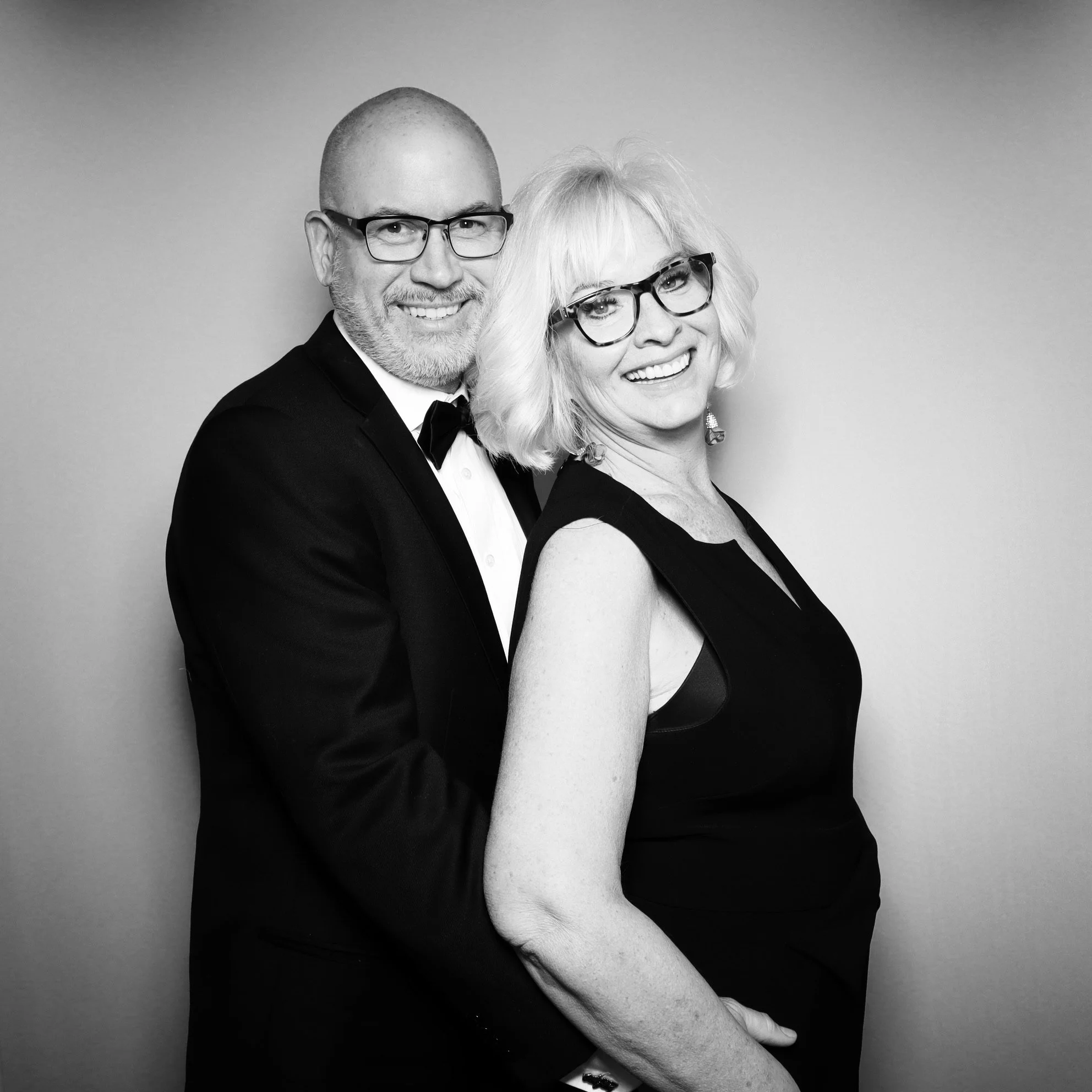 Black and white photo of a smiling man in a tuxedo and a woman in a black dress, both wearing glasses, standing close together against a plain background.