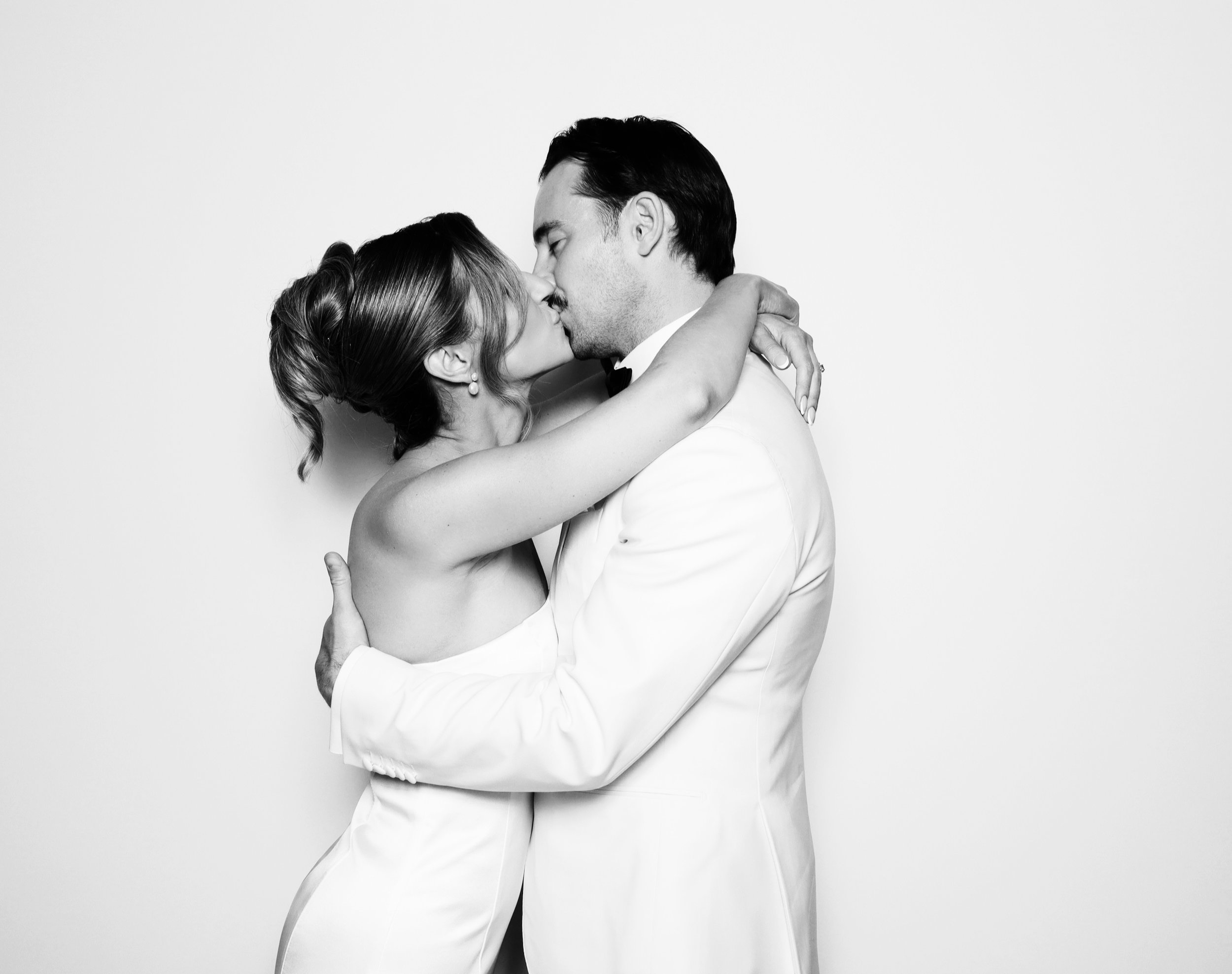 A black and white photo of a couple in wedding attire sharing a kiss, with the woman wearing a strapless dress and the man in a tuxedo.