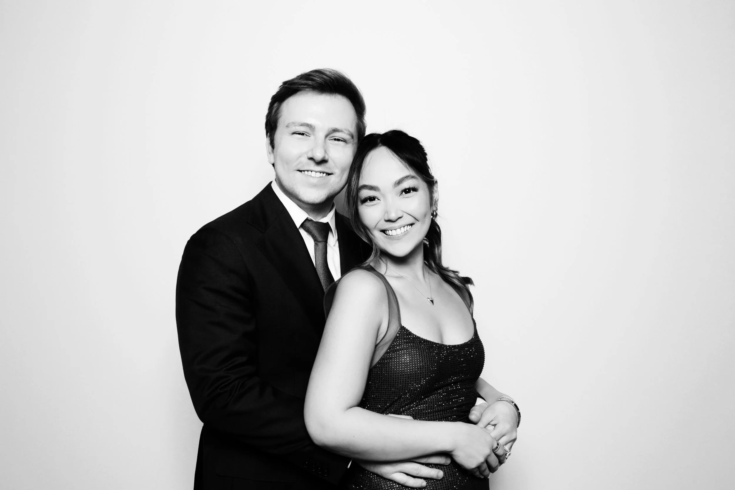 A black and white photo of a smiling man in a suit and a woman in a dress, standing close together against a plain light background.