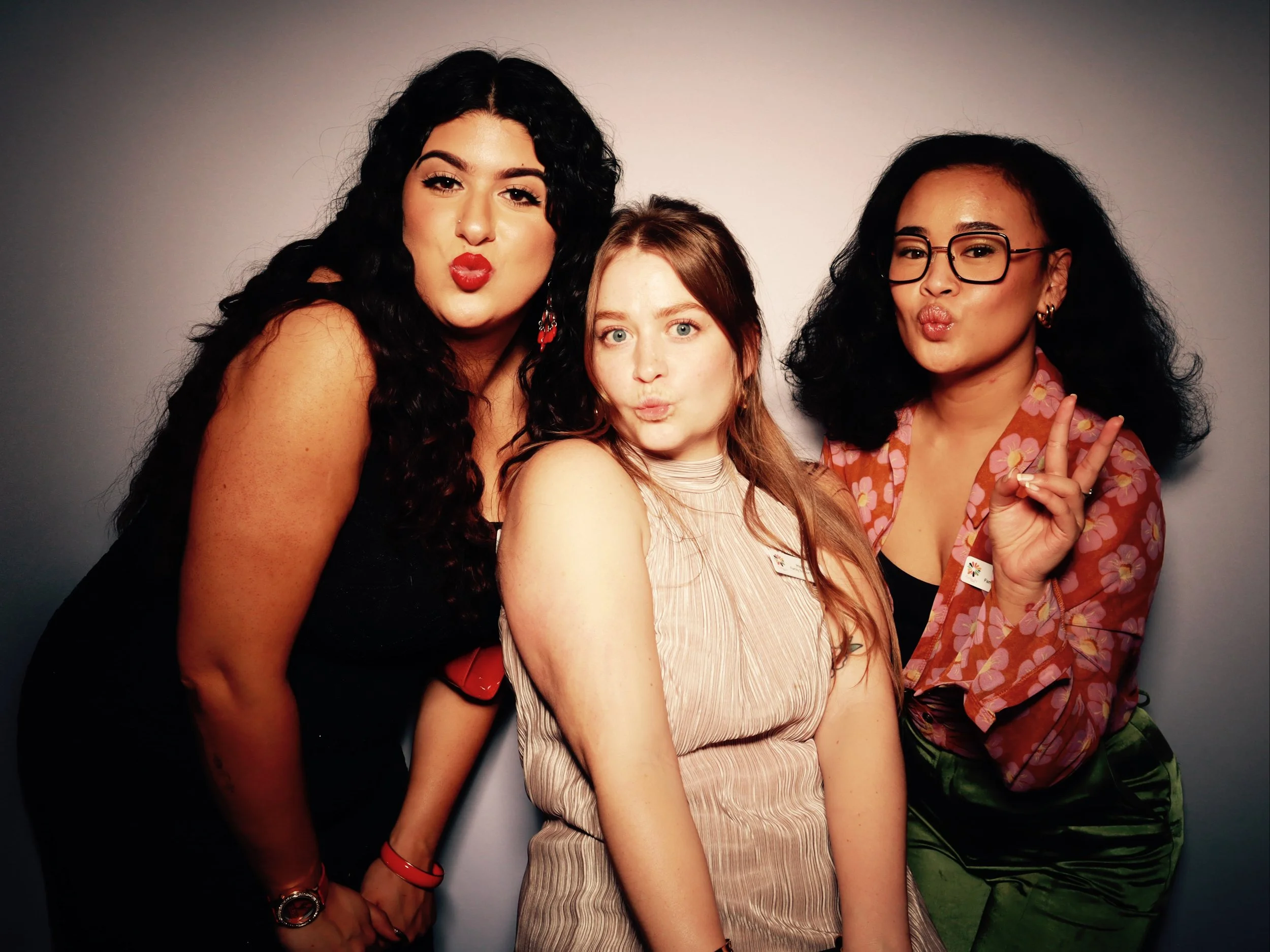 Three women posing together, making kissy faces and a peace sign, against a gray background.