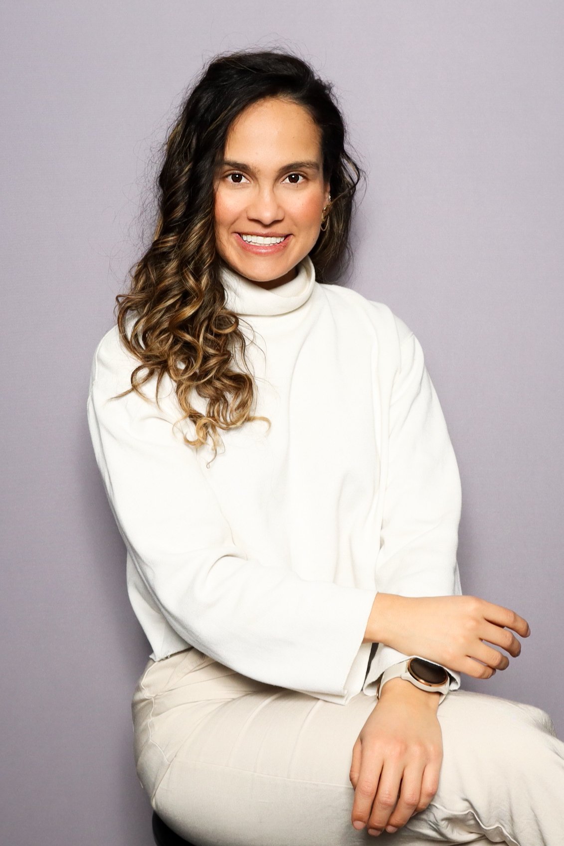 A woman with long wavy brown hair and light skin, smiling, wearing a white turtleneck sweater and white pants, sitting against a light gray background.