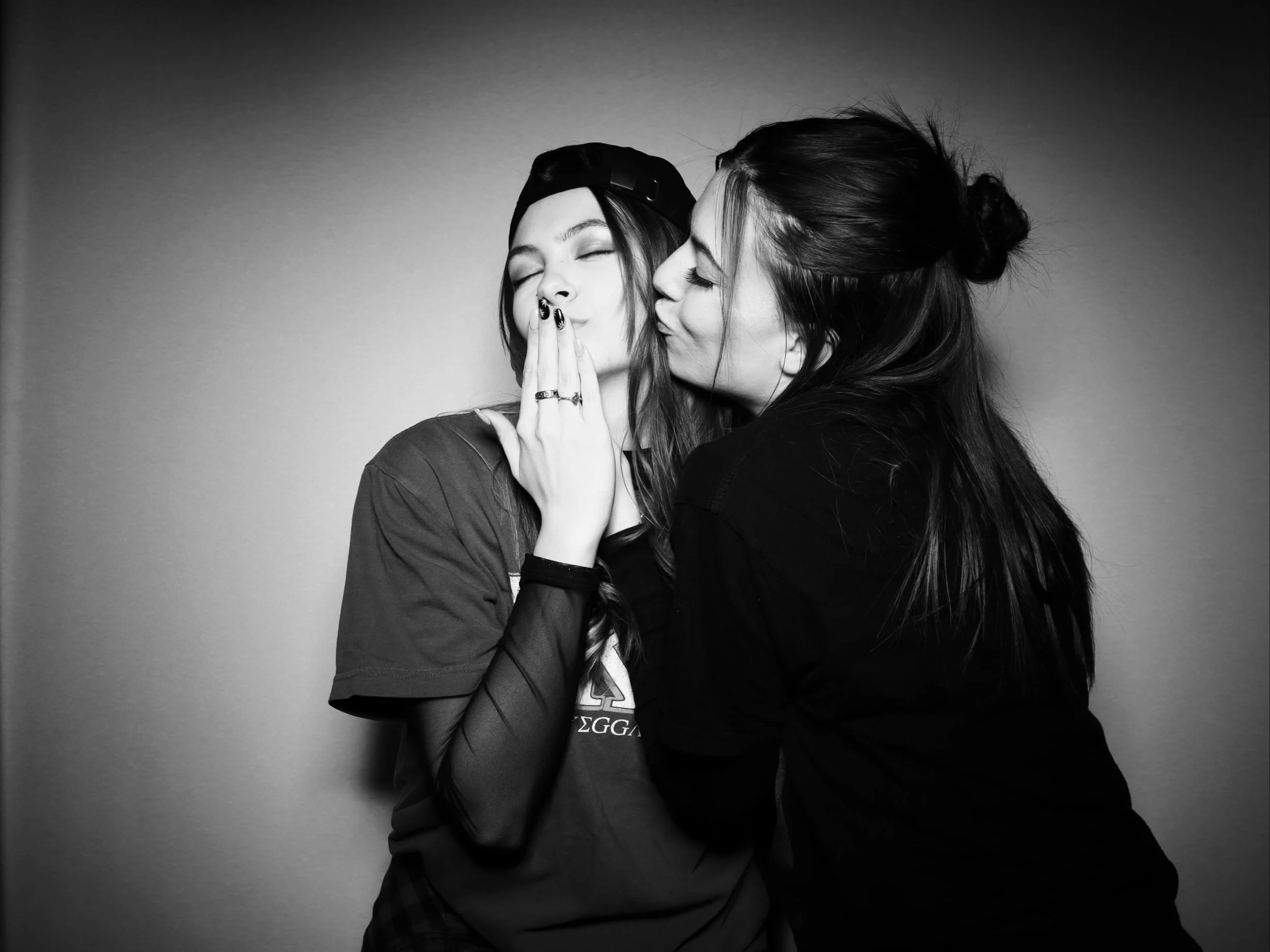 Two young women sharing a kiss on the cheek, one with her eyes closed and the other with her lips puckered, in a black and white photo.
