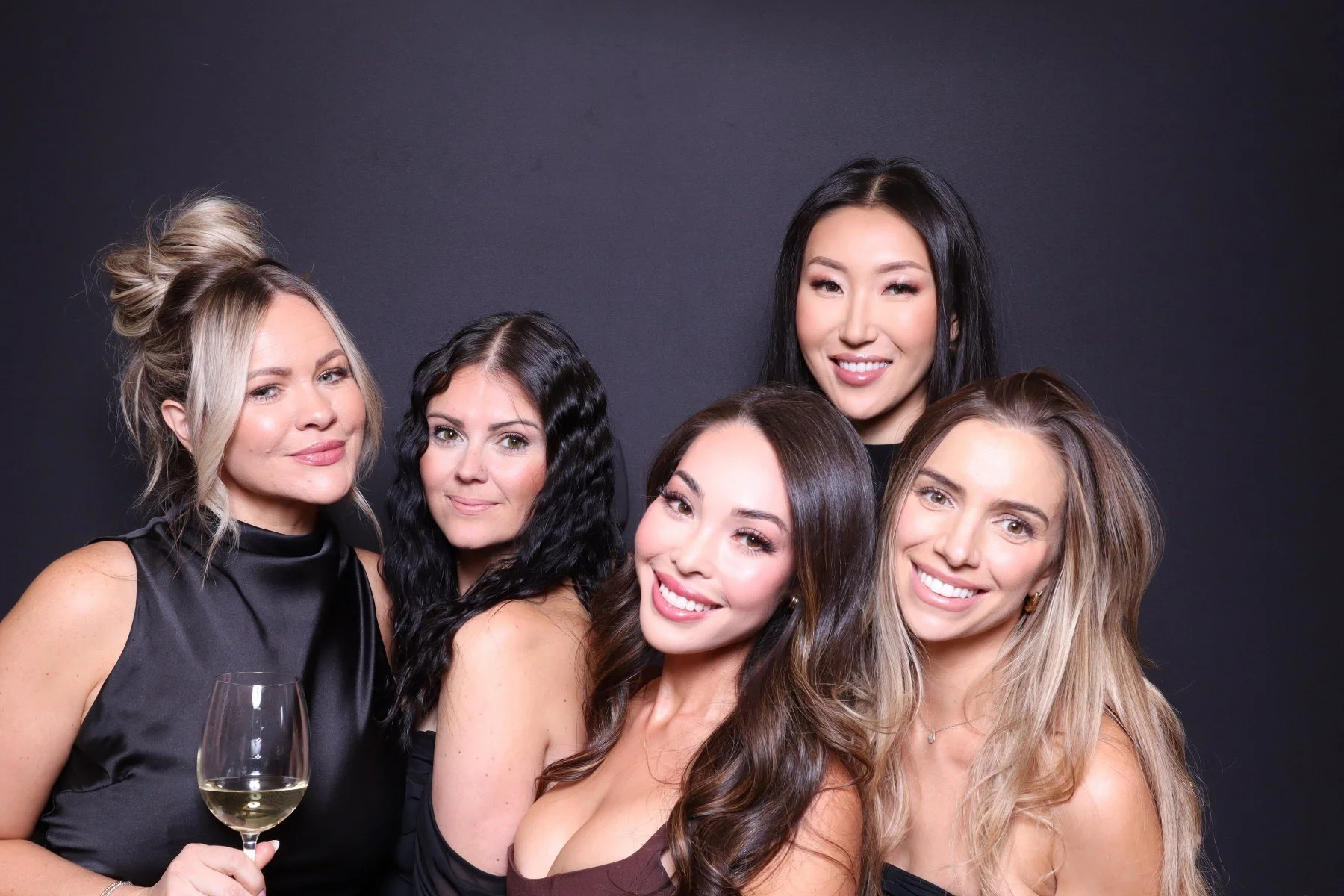 Group of five women at a social gathering, with one woman holding a glass of white wine, posed against a dark background, smiling and looking at the camera.