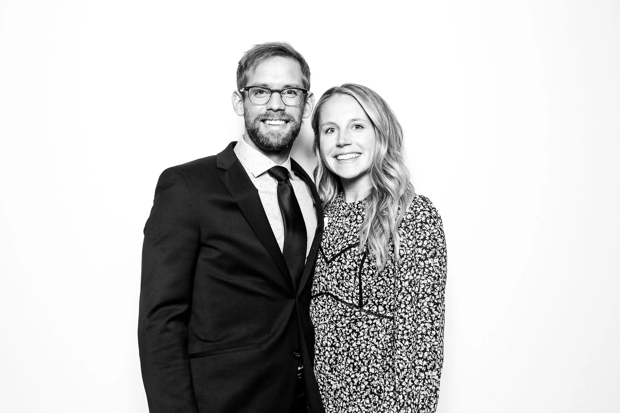 A man and woman smiling and standing close together against a plain white background. The man is wearing glasses, a suit, and a tie. The woman has wavy hair and is wearing a patterned dress.