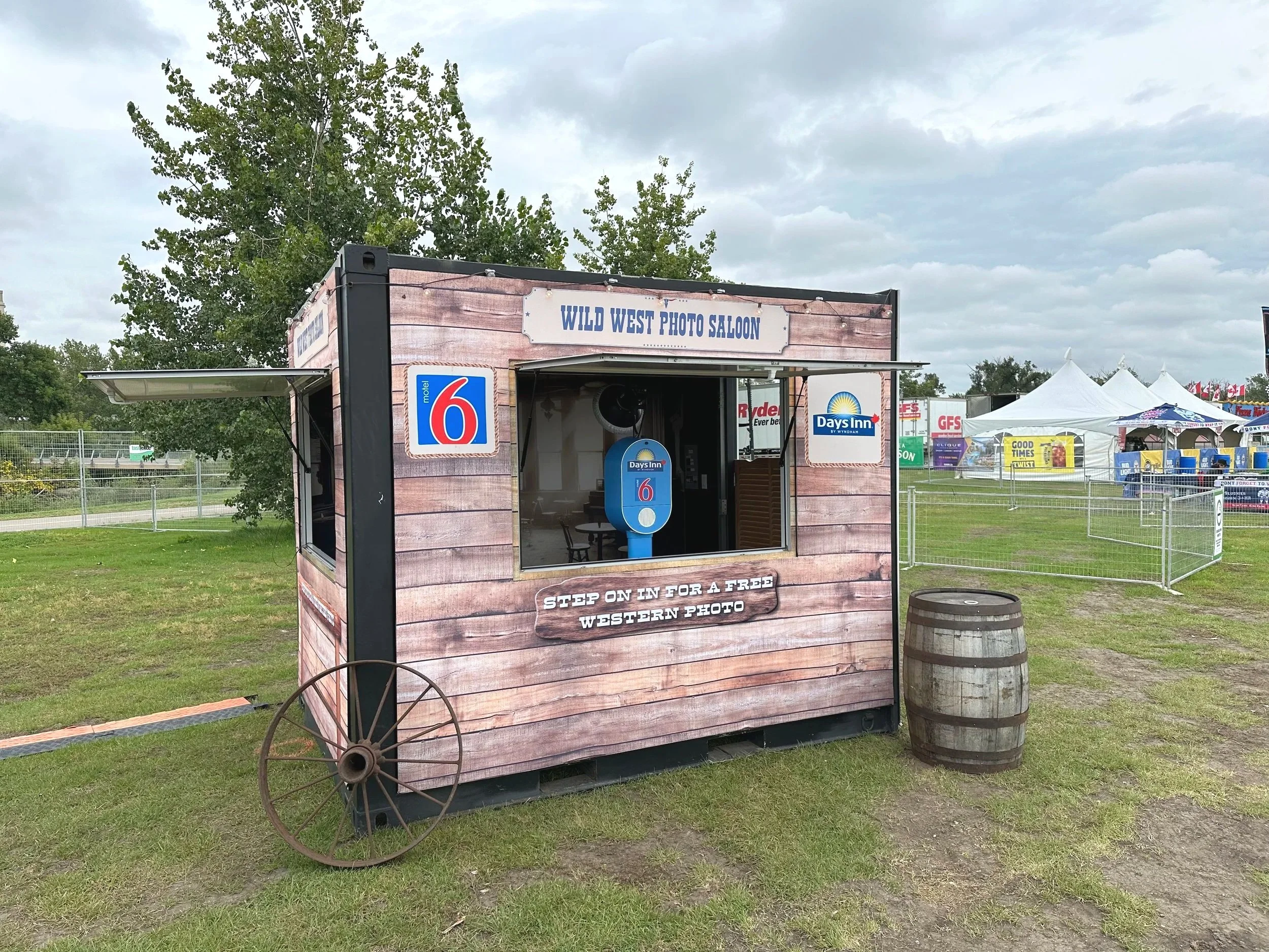 A photo booth styled as a Wild West photo saloon at an outdoor event with tents and carnival rides in the background, featuring a sign that reads 'STEP ON IN FOR A FREE WESTERN PHOTO' and a barrel in front.