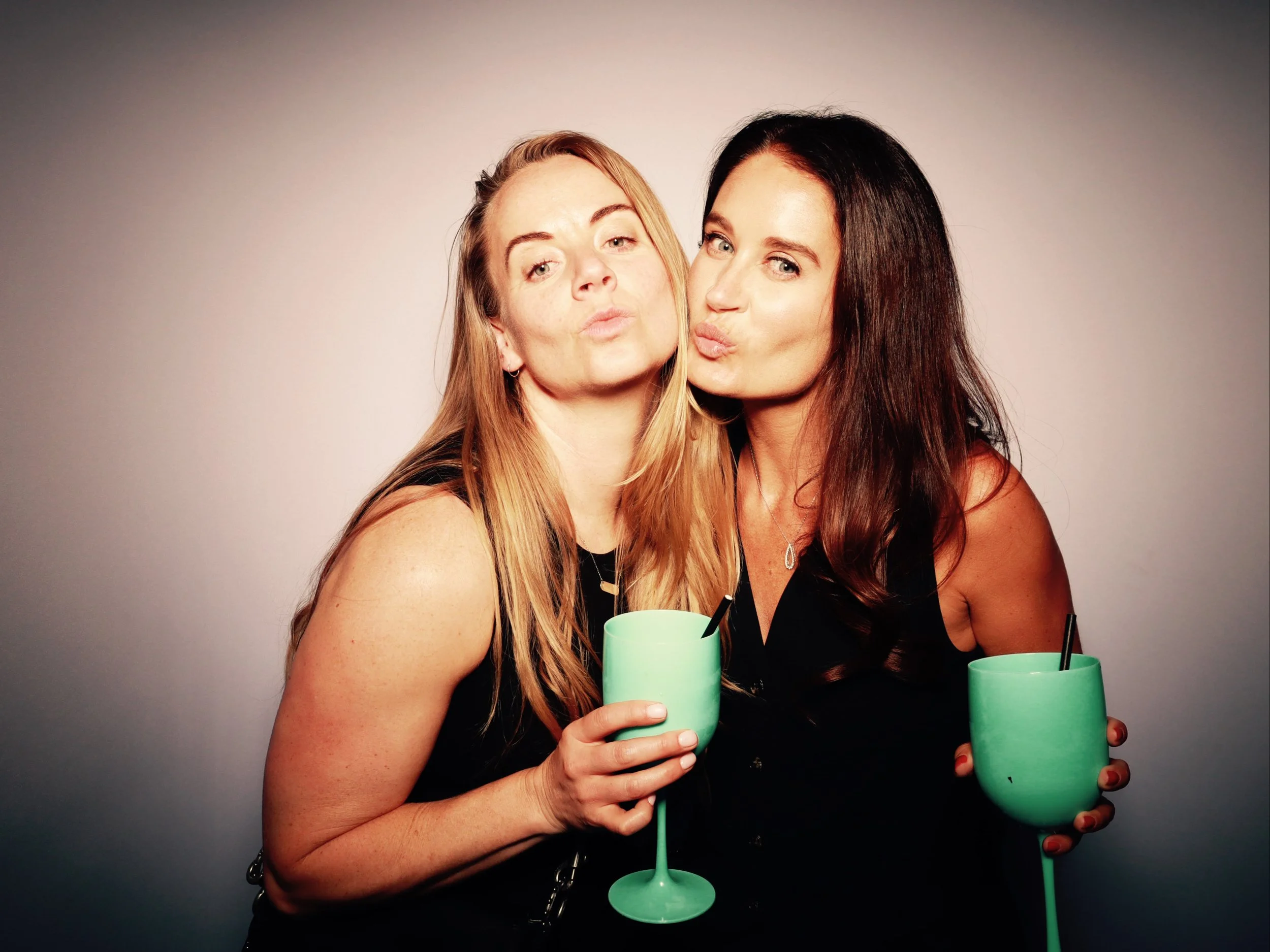 Two women with long hair, one with blonde and one with dark hair, are making kissy faces while holding green glasses with black straws.