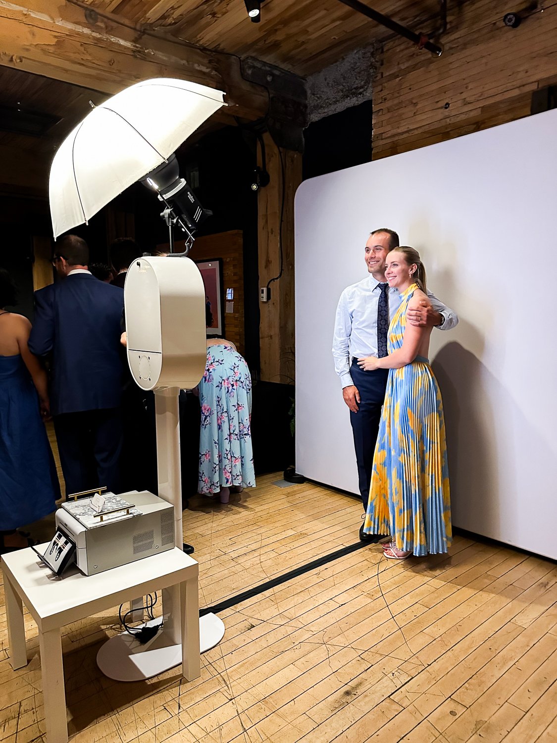 A couple stands together, smiling, in front of a white photo backdrop at a photography session, with a photographer taking their picture. The woman is wearing a colorful, striped dress and the man is in a white shirt and dark pants.