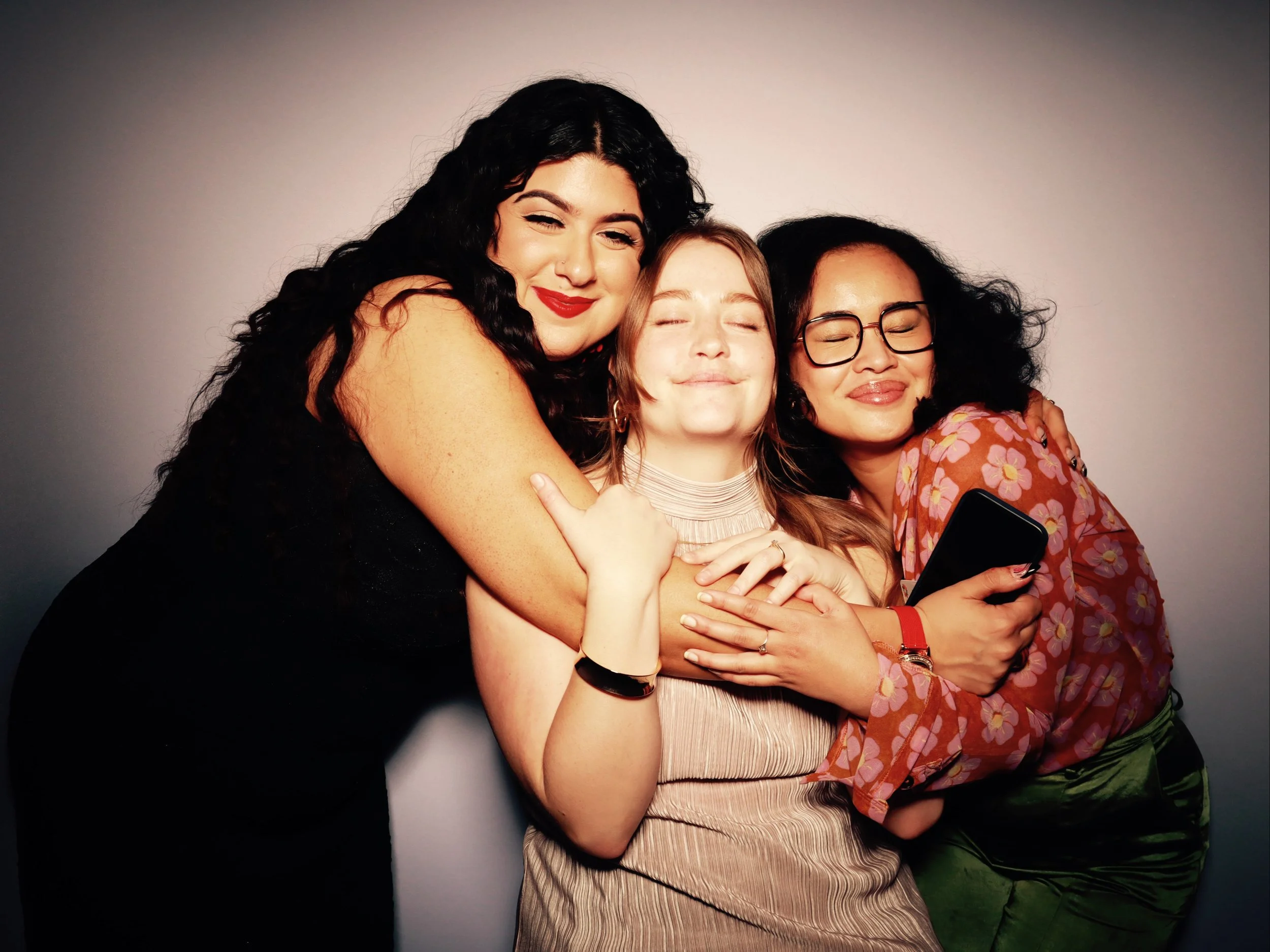 Three women hugging and smiling with arms around each other, one holding a phone, against a plain gray background.